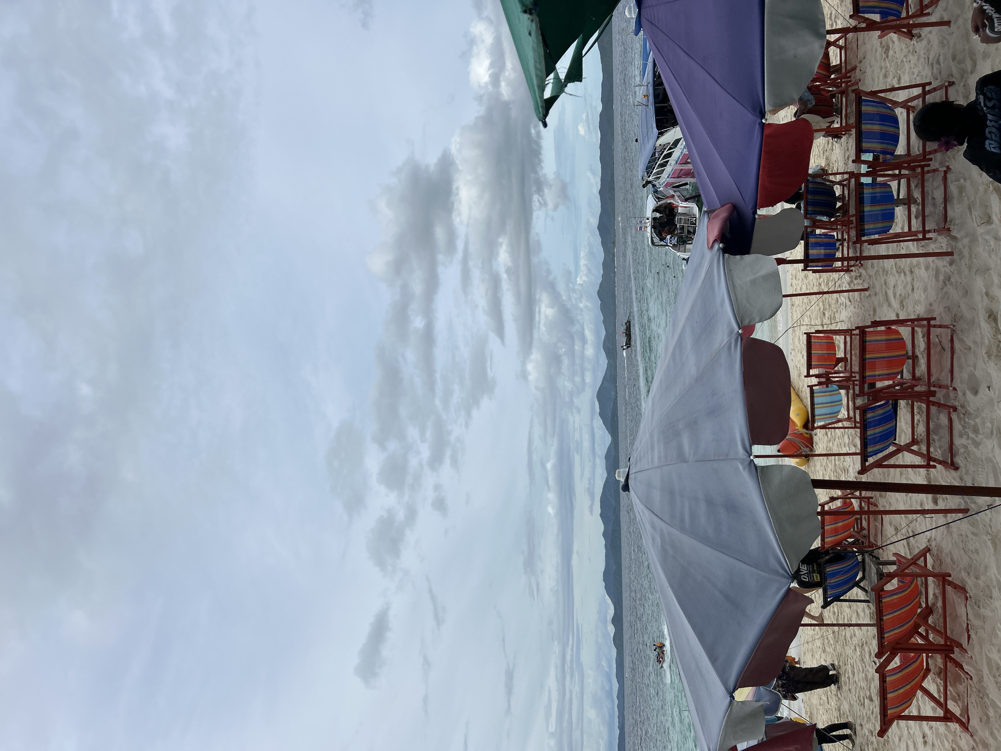 The image depicts a beach scene with several beach chairs and umbrellas set up on a sandy shore. In the background, the ocean is visible with several boats and a distant coastline with mountains under a cloudy sky. The scene appears to be in Tambon Phru Nai, Thailand, suggesting a tropical coastal location. There are a few people visible, some sitting in chairs and others walking. The overall atmosphere seems relaxed, though the cloudy sky might indicate a change in weather.