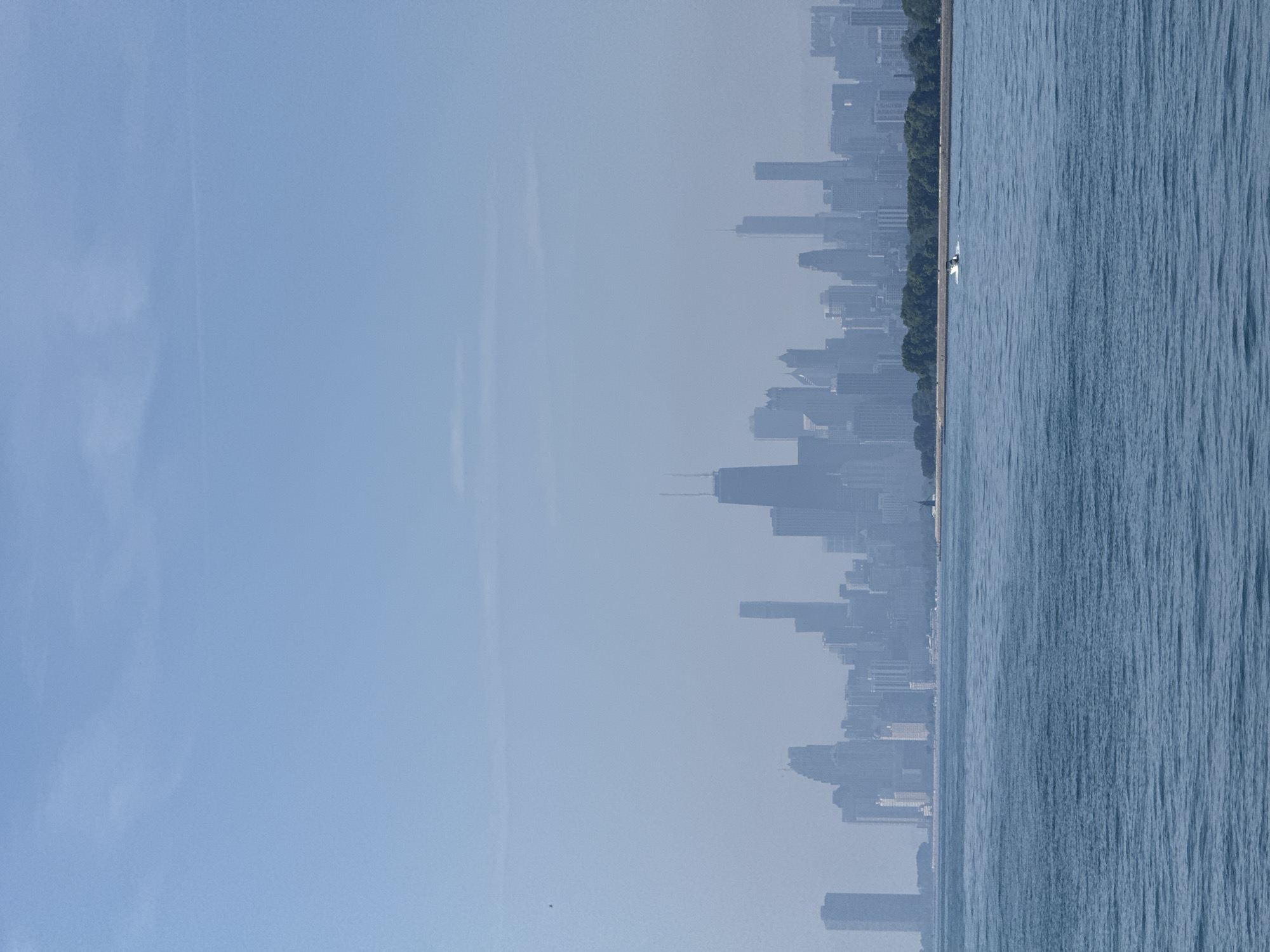 The image captures a hazy view of the Chicago skyline from across Lake Michigan. The city's iconic skyscrapers, including the John Hancock Center and the Willis Tower, are silhouetted against a pale blue sky. The water in the foreground is a deep blue, with gentle ripples suggesting a calm day. A small white boat is visible in the distance, traversing the lake. Trees line the shoreline in front of the city, adding a touch of green to the urban landscape. The overall impression is one of a serene day in Chicago, with the city appearing as a distant, almost ethereal presence across the vast expanse of the lake.