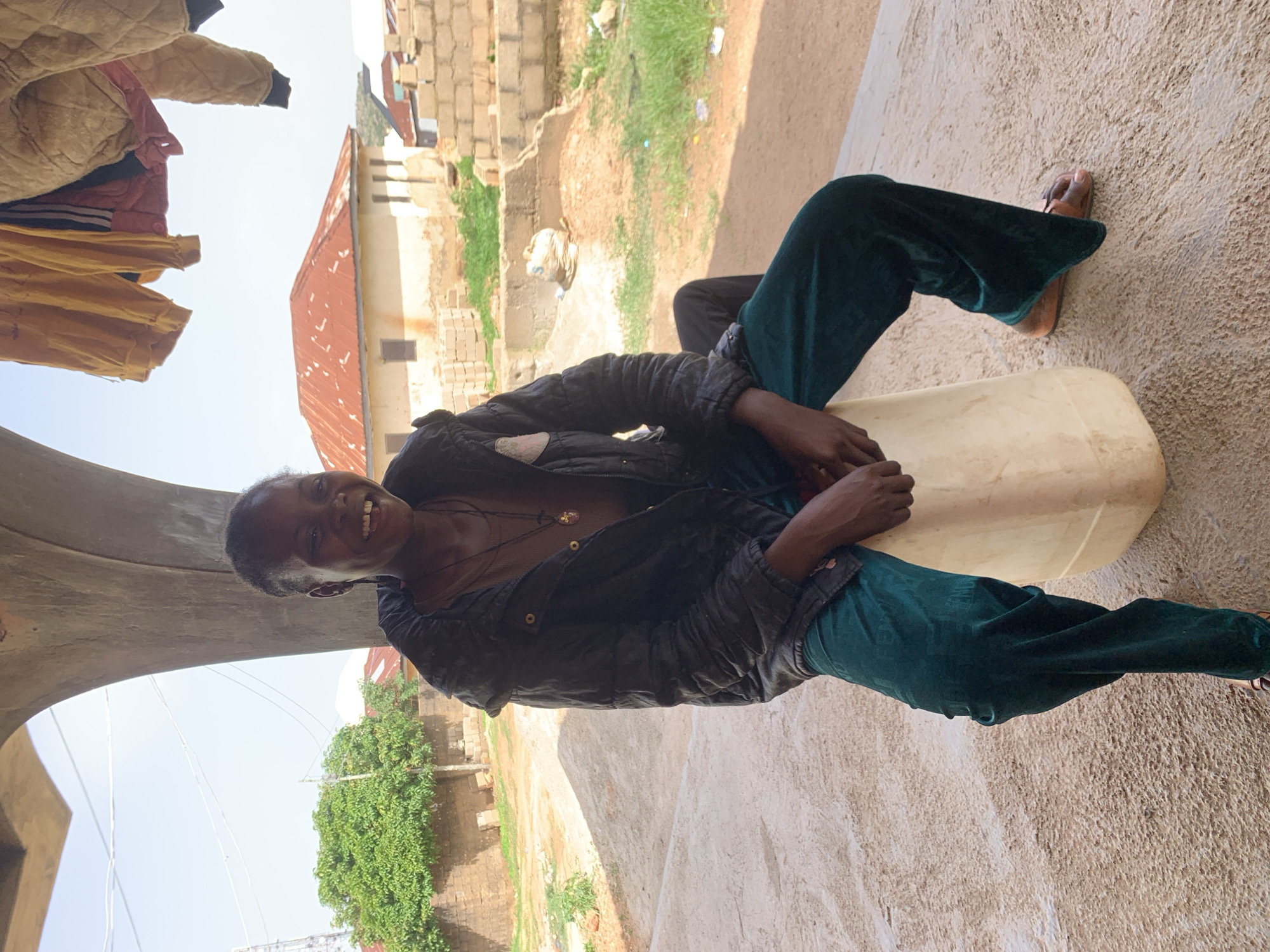 A Black woman in a black puffer jacket and teal velvet pants is seated on a large, off-white plastic container. She is smiling broadly and looking towards the viewer, with her head tilted slightly to her left. She is wearing a necklace with a pendant. The background shows a rural or semi-rural setting, possibly on the outskirts of Jos, Nigeria. There are buildings with red corrugated roofs, some brick construction, and greenery. Clothing hangs on a line in the upper right of the frame. The ground is a mix of sand and concrete.