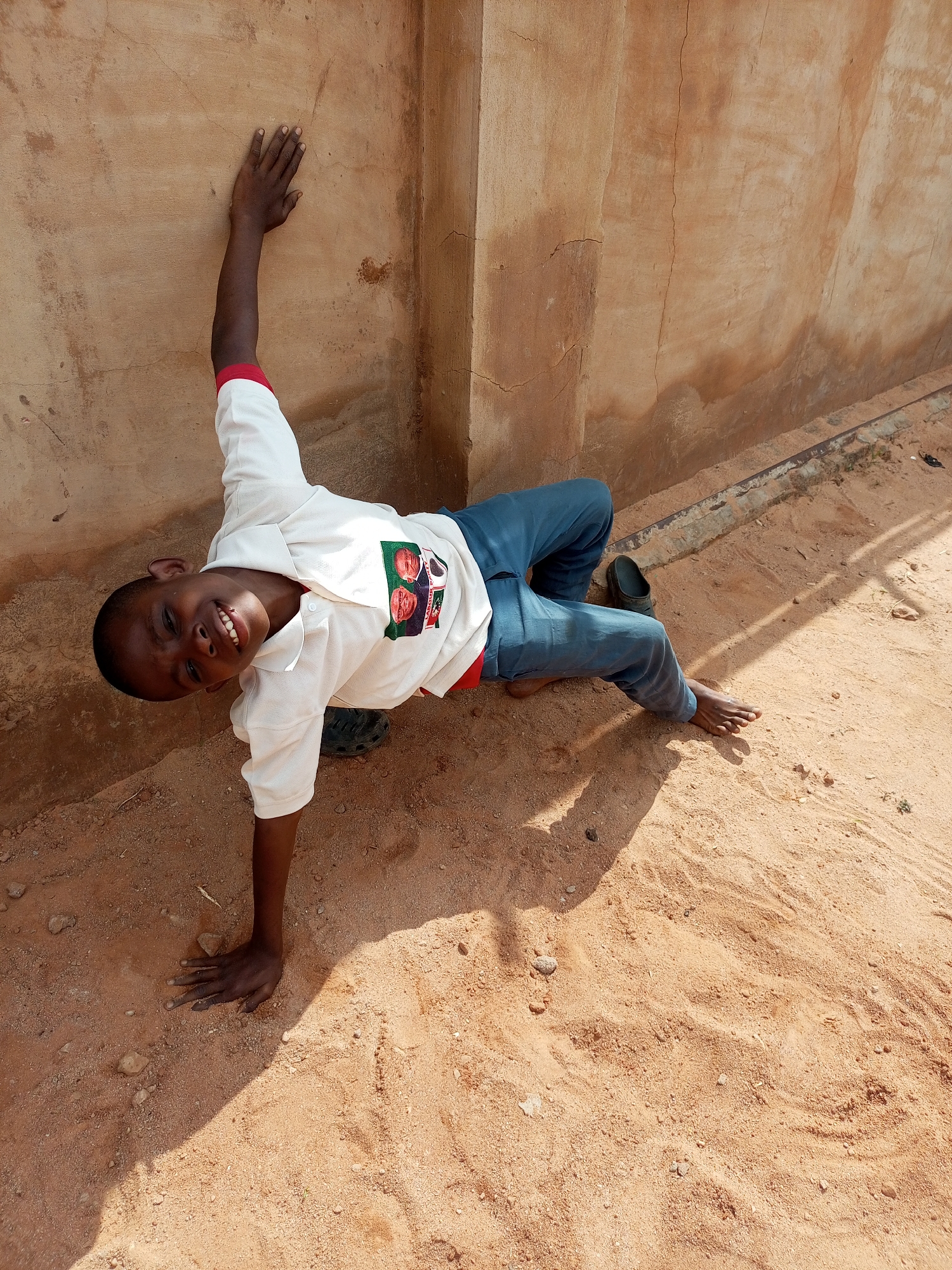 In Tempure, Nigeria, a young boy, appearing to be around 6-8 years old, is captured in a playful pose outdoors. He is dressed in a white collared shirt with red trim on the sleeves and a red undershirt. He wears blue trousers and one dark shoe is visible on the ground near his left leg. The boy is leaning against a textured, light brown wall that shows signs of age and wear, including cracks and discoloration. He is supported by his left arm, which is extended upwards and placed flat against the wall, while his right hand is on the sandy ground for balance. His body is angled, and he is looking towards the camera with a wide, joyful smile. The ground beneath him is composed of sand and small pebbles, with a distinct shadow cast by the wall and possibly an unseen object or structure. The lighting suggests it is daytime, with a clear sky.