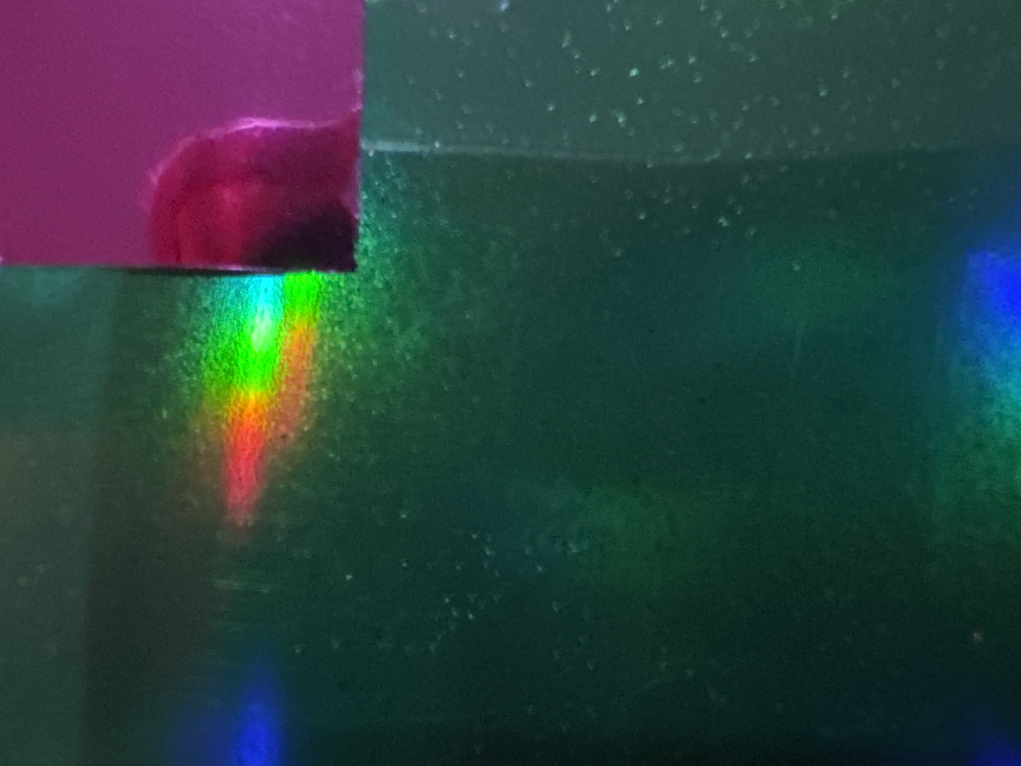 The image is a close-up, abstract shot of a holographic surface reflecting light. There is a distinct rainbow-like spectrum of colors, including red, orange, yellow, green, and blue, appearing as a diffused band across the dark green, textured surface. To the right, a bright pink or magenta surface is visible, with a blurred, red, fuzzy object or texture on it. The image does not depict any recognizable landmarks or scenes from Waterford, Ireland, and therefore cannot be geolocated there. It is an abstract representation of light and surface texture.