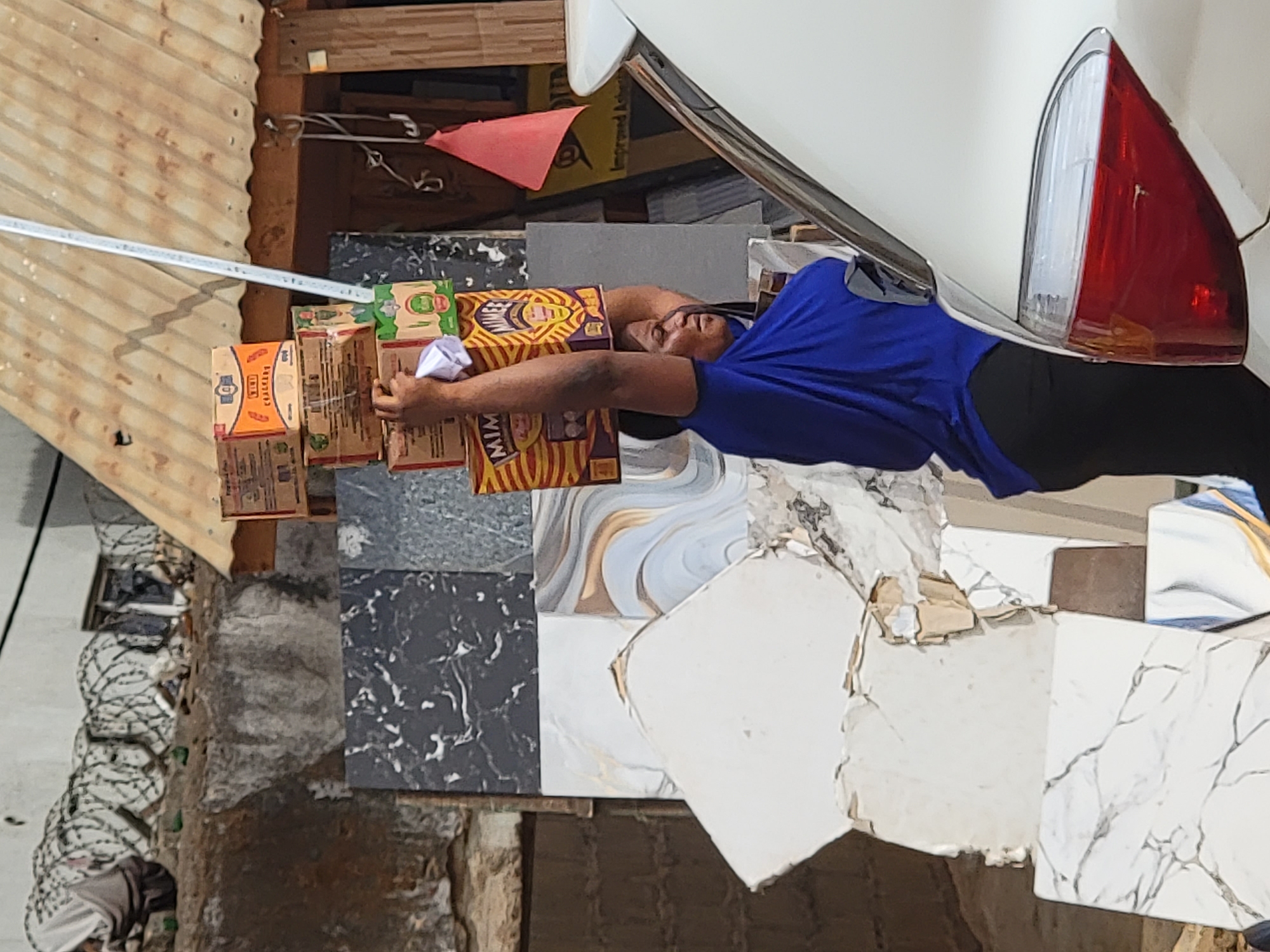 A man in a blue shirt and black pants lies horizontally across a collection of cardboard boxes and what appears to be broken tiles near a white car with a red and white taillight. The boxes appear to contain food items. The surrounding environment includes a corrugated metal roof, a structure made of wooden beams, and what appears to be a street or alleyway. The location is identified as Maiburiji, Nigeria.