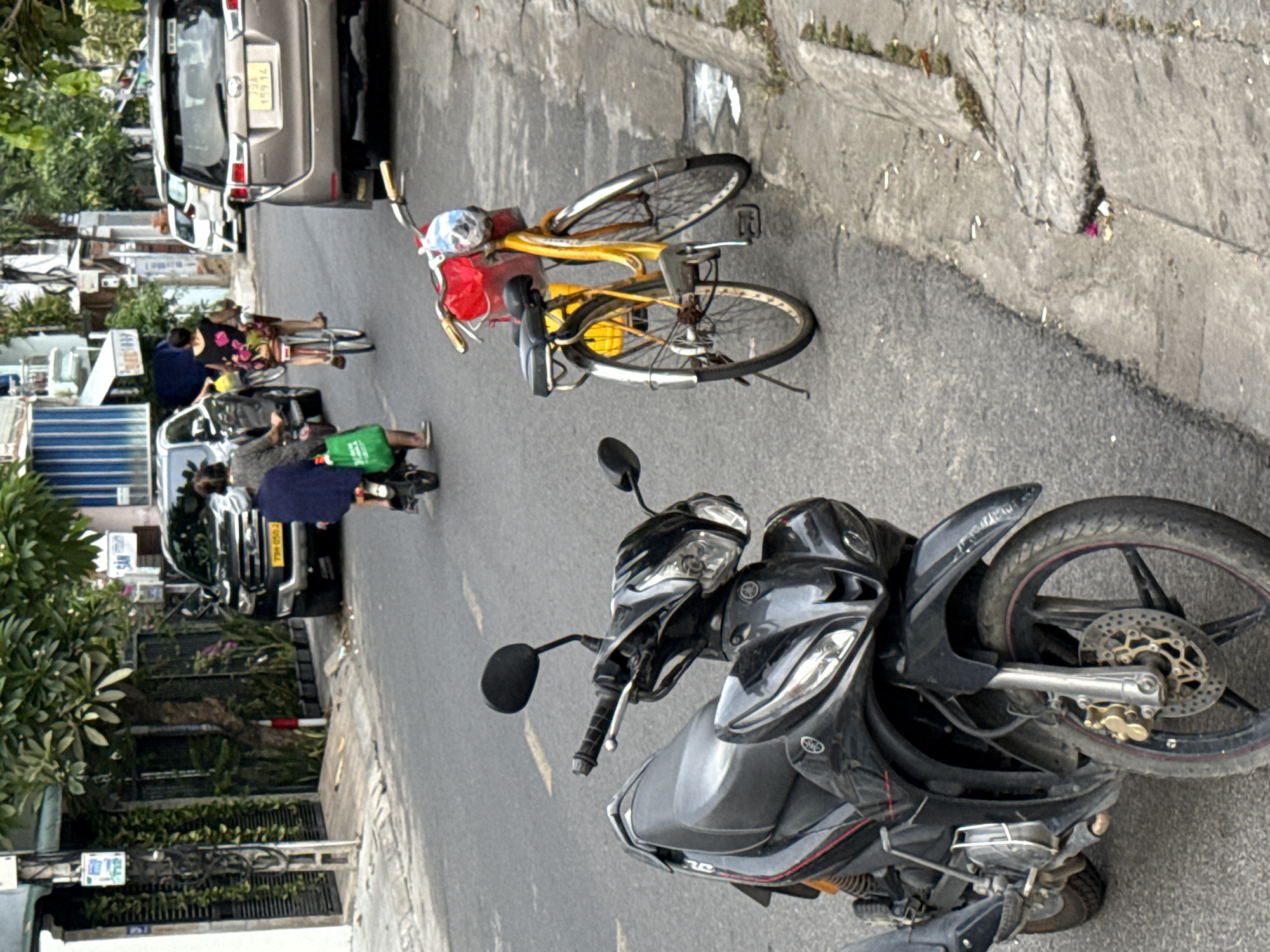 In a city street scene in Nha Trang, Vietnam, a black motorcycle is parked on the left side of the frame, facing away from the viewer. The motorcycle has a black seat, a prominent headlight assembly, and silver accents on its body. To the right of the motorcycle, a yellow bicycle is parked, facing towards the viewer, with a red basket containing a clear plastic bag on its handlebars. The bicycle has black tires and a black seat.
In the background, a grey SUV is parked on the right side of the street, facing away from the viewer. Its license plate is visible and reads "79A 159.14". Further down the street, a black SUV is parked on the left side, also facing away from the viewer. A person on a smaller motorcycle is seen to the left of the black SUV, wearing a dark blue shirt and carrying a green bag. To the right of the black SUV, a person on a bicycle is seen wearing a black dress with a floral pattern, also facing away from the viewer. Trees and buildings line the street. The road is asphalt, and a concrete sidewalk is visible on the right side, with some litter and vegetation. The lighting suggests it is daytime.