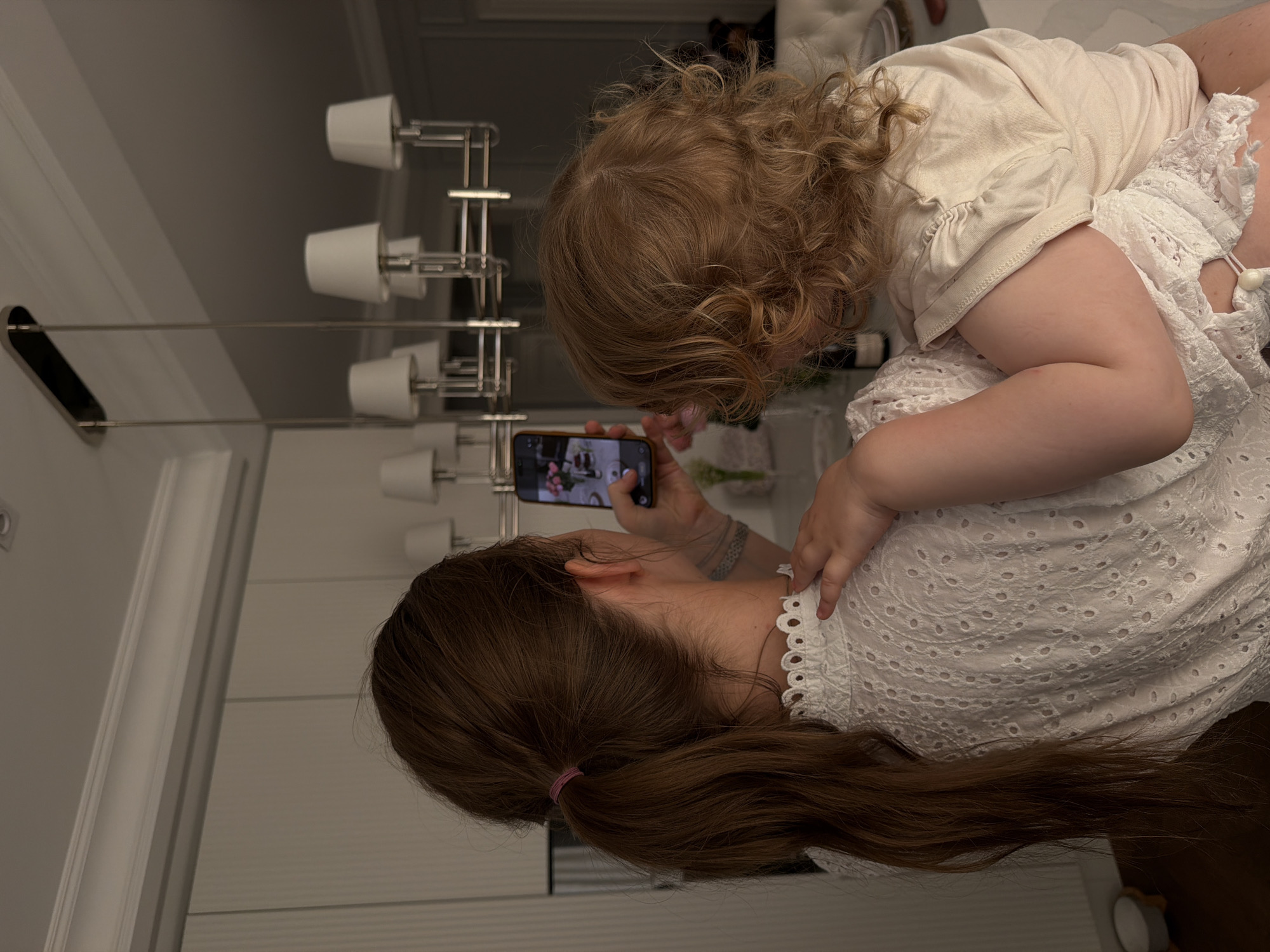 A woman is holding a smartphone while a young child leans on her back. The woman has long brown hair tied back with a pink hair tie, and she is wearing a white, lace-patterned top. The child, who has curly blonde hair, is wearing a beige shirt and a white dress. The woman is holding her phone with both hands and appears to be taking a photo of something. The scene is indoors, with a multi-bulb light fixture visible in the background. The location could be a home or apartment in Moscow, Russia, but there are no distinctive landmarks or identifiers to confirm this.