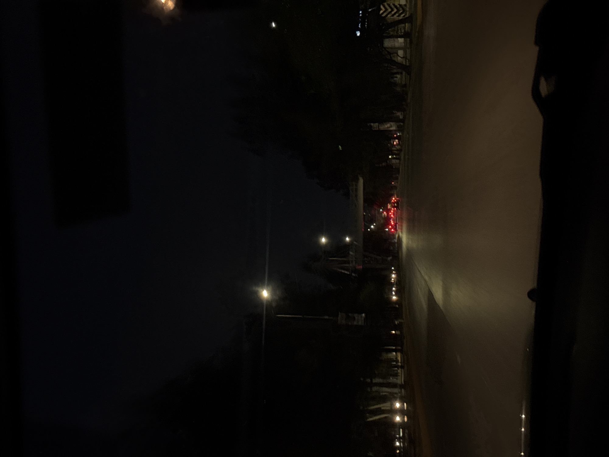 The image depicts a night scene in San Bernardino Tlaxcalancingo, Mexico. The view is likely taken from inside a vehicle looking out onto a road illuminated by streetlights and the headlights and taillights of distant vehicles. The road surface appears smooth and reflects some of the light. The surrounding environment is dark, with trees and buildings only faintly discernible against the night sky. The right edge of the frame is occupied by the interior of the vehicle.