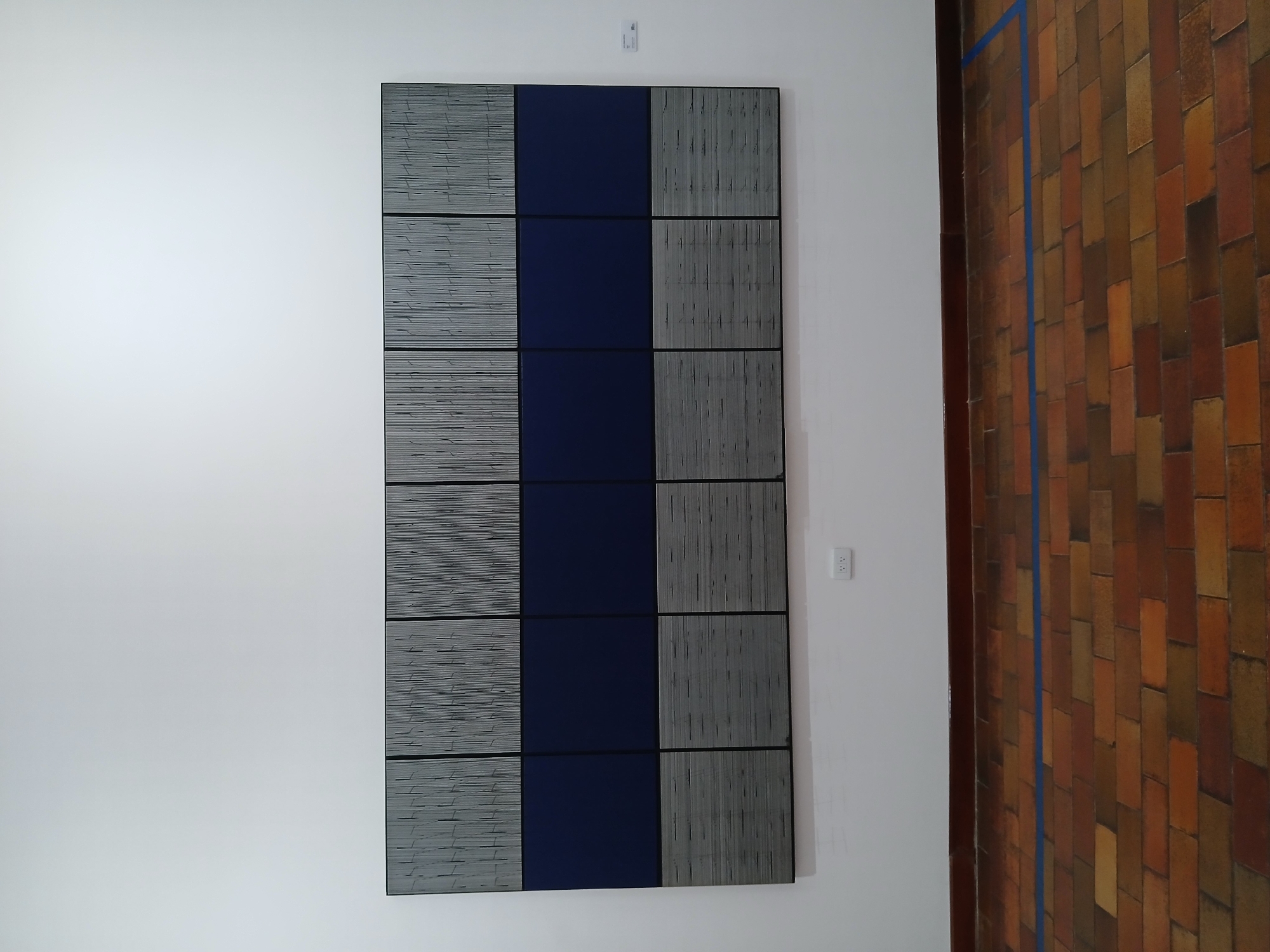 The image shows a rectangular artwork hanging on a white wall. The artwork consists of nine square panels arranged in a 3x3 grid. The panels on the left and right columns are gray with horizontal or vertical striations, while the panels in the center column are solid blue. The panels are separated by thin black lines, creating a grid-like pattern. Above the artwork is a small white object, possibly a sensor. To the right of the artwork, a wall with brown, rectangular tiles is visible, marked with a vertical blue line near the edge. A white electrical outlet is positioned on the wall to the right of the artwork. The location is Unknown, Unknown.