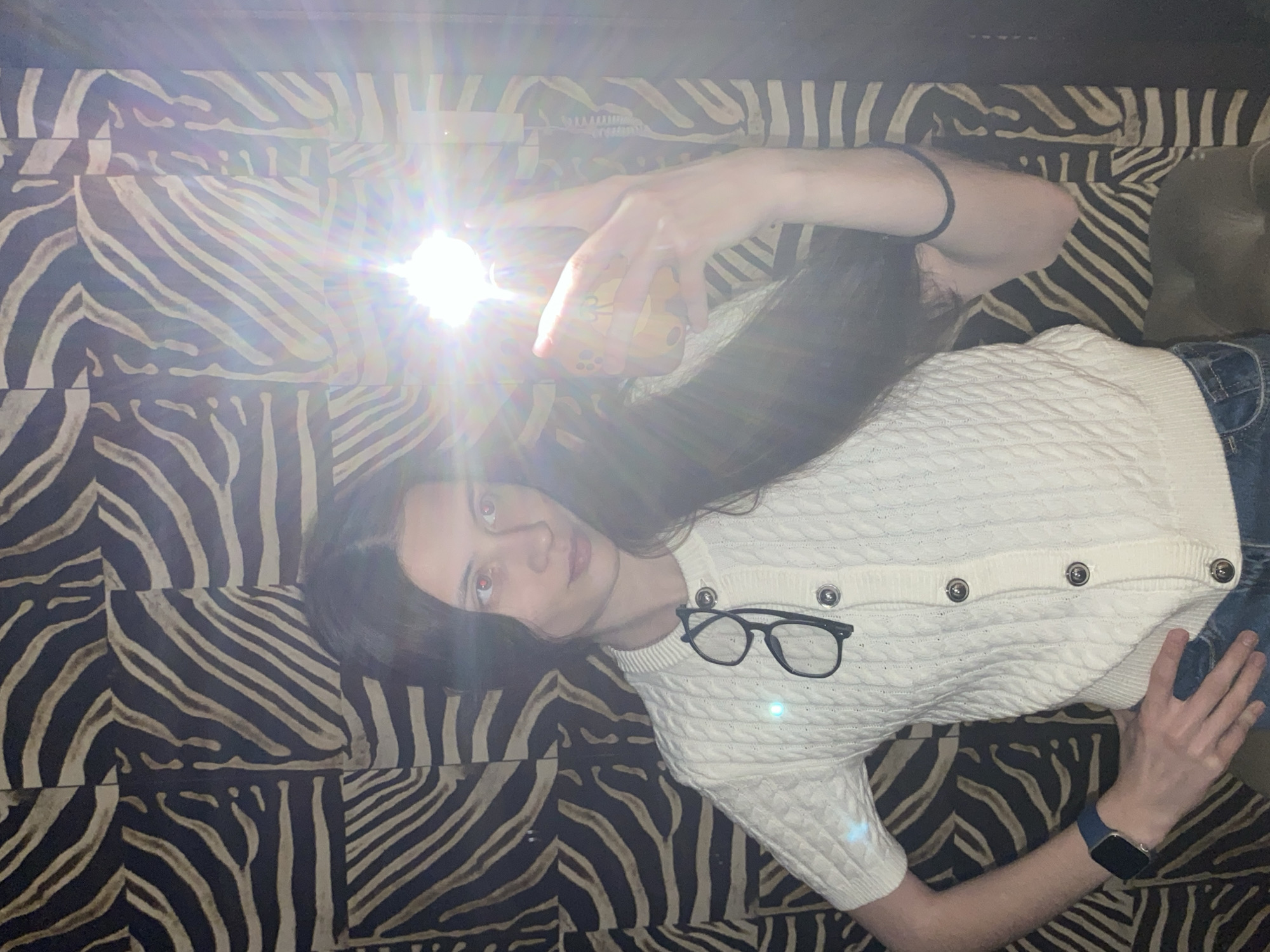 A person, appearing to be female, is lying down on a surface with a distinct black and off-white zebra-print pattern, which seems to be composed of square or rectangular sections. The scene is indoors in Votkinsk, Russia.
The individual has long, dark hair spread out beneath her head. She is wearing a white cable-knit short-sleeved cardigan with silver-toned buttons and blue jeans. A pair of black-framed eyeglasses rests on her cardigan. On her left wrist, she wears a blue smartwatch and a black elastic hair tie. Her right hand holds a smartphone directly towards the camera, emitting a very bright flash that causes significant lens flare and illuminates her face.
The primary activity is the person taking a selfie. The intense flash suggests the photograph was taken in low-light conditions, possibly at night. Her eyes show a "red-eye" effect due to the direct flash. Her expression is neutral as she looks towards the camera. There is no visible text in the image.