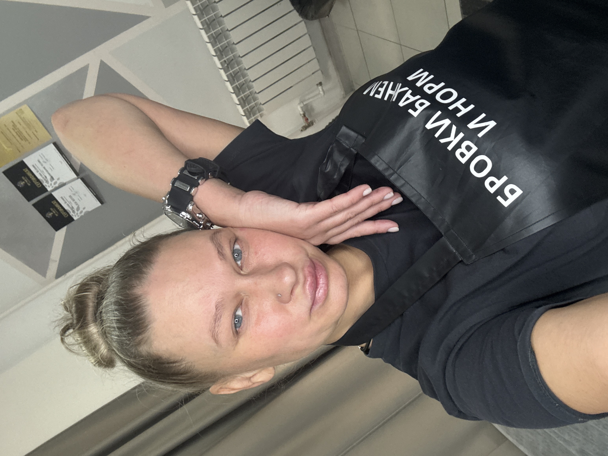 A woman is captured in a selfie-style portrait, slightly angled, in what appears to be an indoor professional setting. She has fair skin, blue eyes, and light-colored hair pulled back into a bun. A subtle nose piercing is visible. She is wearing a black t-shirt and a black apron or smock with prominent white Cyrillic text. On her left wrist, she wears a robust, dark-colored watch, likely a sports watch. Her right hand is raised, resting gently on her head, showcasing neatly manicured nails. Her facial expression is calm and neutral.
The white text on her black apron, which is inverted in the image, reads "БРОВКИ ВАЖНЕЙ И НОРМ." This phrase translates to "Eyebrows are more important and normal" or "Eyebrows are important and the norm," strongly suggesting she is a beauty professional specializing in eyebrow services.
The background reveals a grey wall with a light geometric pattern. To her left, several certificates or awards are displayed, including one gold-colored and multiple black-and-white ones, one of which clearly reads "СЕРТИФИКАТ" (CERTIFICATE). To her right, a white radiator or heating unit is visible, along with dark, possibly brown or grey, curtains. The overall ambiance indicates an indoor, possibly commercial, space such as a beauty salon or studio, located in Perm, Russia, as per the provided context.