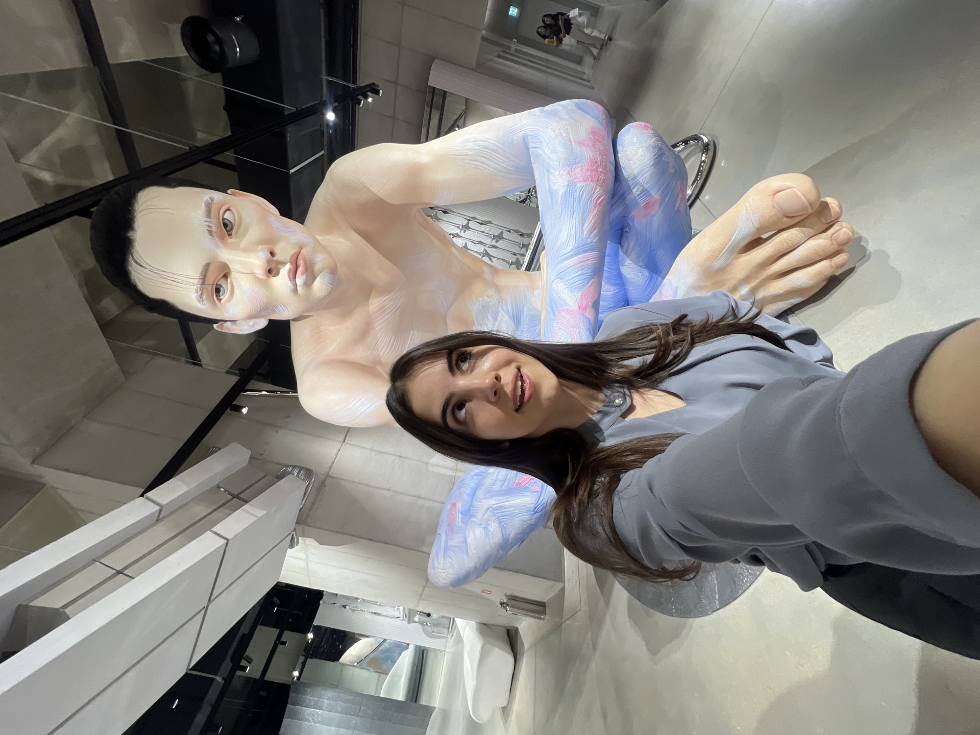 A young woman is captured in a selfie taken indoors, likely within an art gallery or museum in Seoul, South Korea. The image is rotated counter-clockwise, showing her from the chest up, looking slightly upwards and to her right with a subtle smile. She has long, dark hair and is wearing a plain grey top.
Dominating the scene behind and to the side of her is a large, light-skinned, human-like sculpture. This stylized figure has dark hair pulled back, light-colored eyes, and a serene expression. Its skin on the torso and face is a pale, realistic tone, while its limbs and parts of its body are adorned with artistic blue and pink brushstrokes, giving it a painted or ethereal appearance. The sculpture appears to be in a reclined or seated position, with its legs bent. Part of a metallic chair or support structure for the sculpture is visible.
The background features modern architectural elements, including minimalist light grey walls, dark structural beams, and reflective surfaces, possibly mirrors, which show inverted reflections of the space and other distant figures. Light fixtures are visible on the ceiling. The overall setting suggests a contemporary and well-lit exhibition space. There is no visible text in the image.