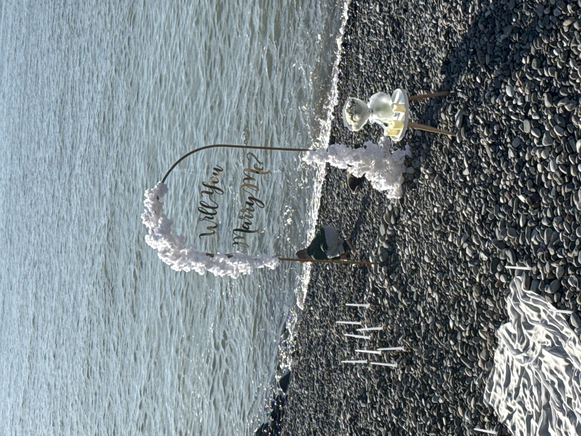 The scene is a "Will You Marry Me?" proposal setup on a rocky beach, likely in Batumi, Georgia, as it is a coastal city with pebble beaches. An arched sign with "Will You Marry Me?" inscribed on it is decorated with white faux flowers. A mannequin head is placed on a stand adjacent to the arch. Several white poles, perhaps to hold candles, are arranged in a pattern on the beach. The Black Sea is visible in the background. The location appears to be prepared for a staged proposal.