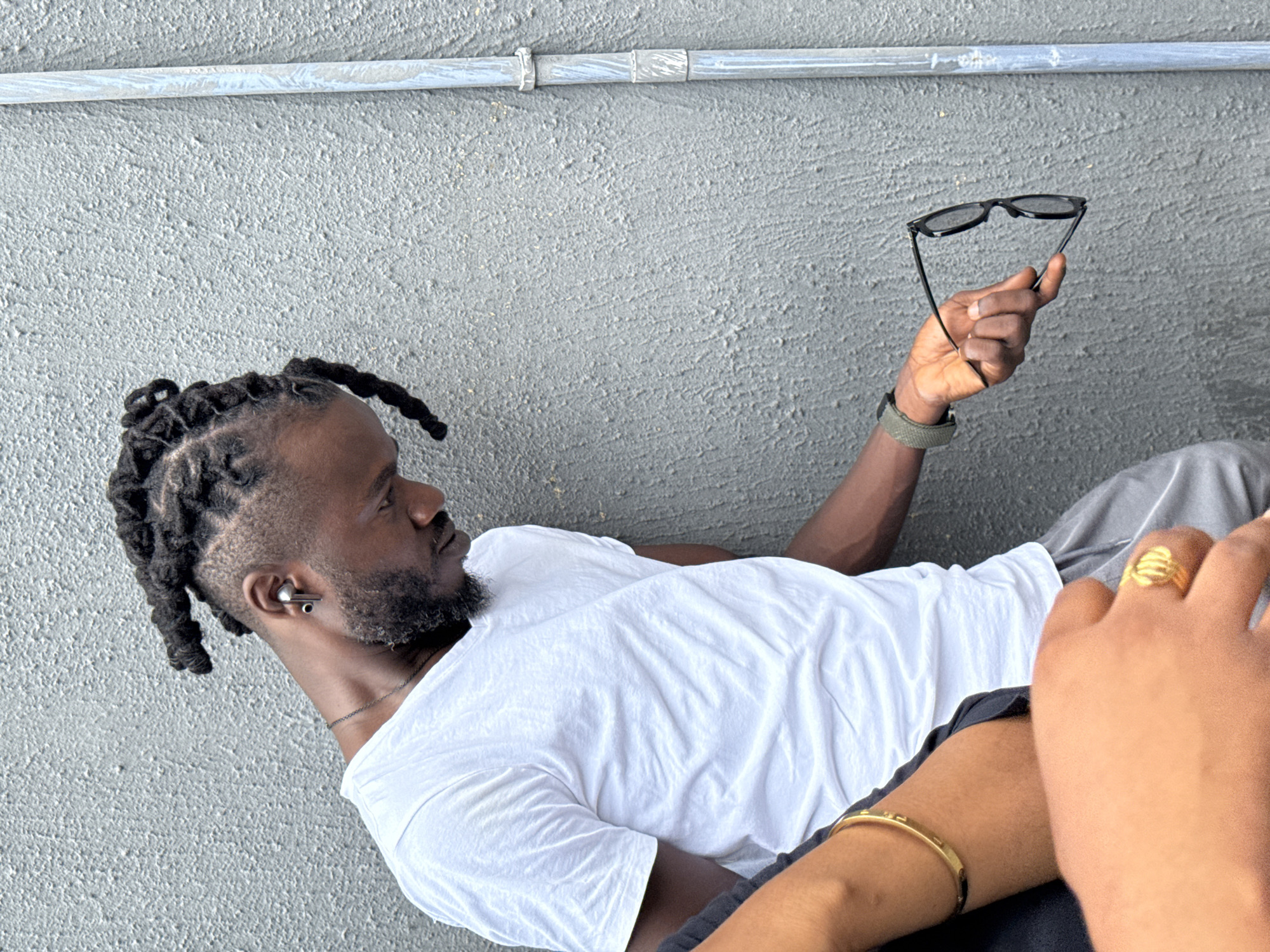 The image depicts a Black man with dreadlocks and a beard, wearing a white t-shirt, lying on what appears to be a gray concrete surface. He is holding a pair of black sunglasses in his right hand. A metal pipe runs horizontally across the top of the frame. In the foreground, partially obscuring the man, is another person's hand with a gold ring and a gold bracelet on their arm. This could be located in the city of Ojomu, Nigeria, but that cannot be determined by the image alone.