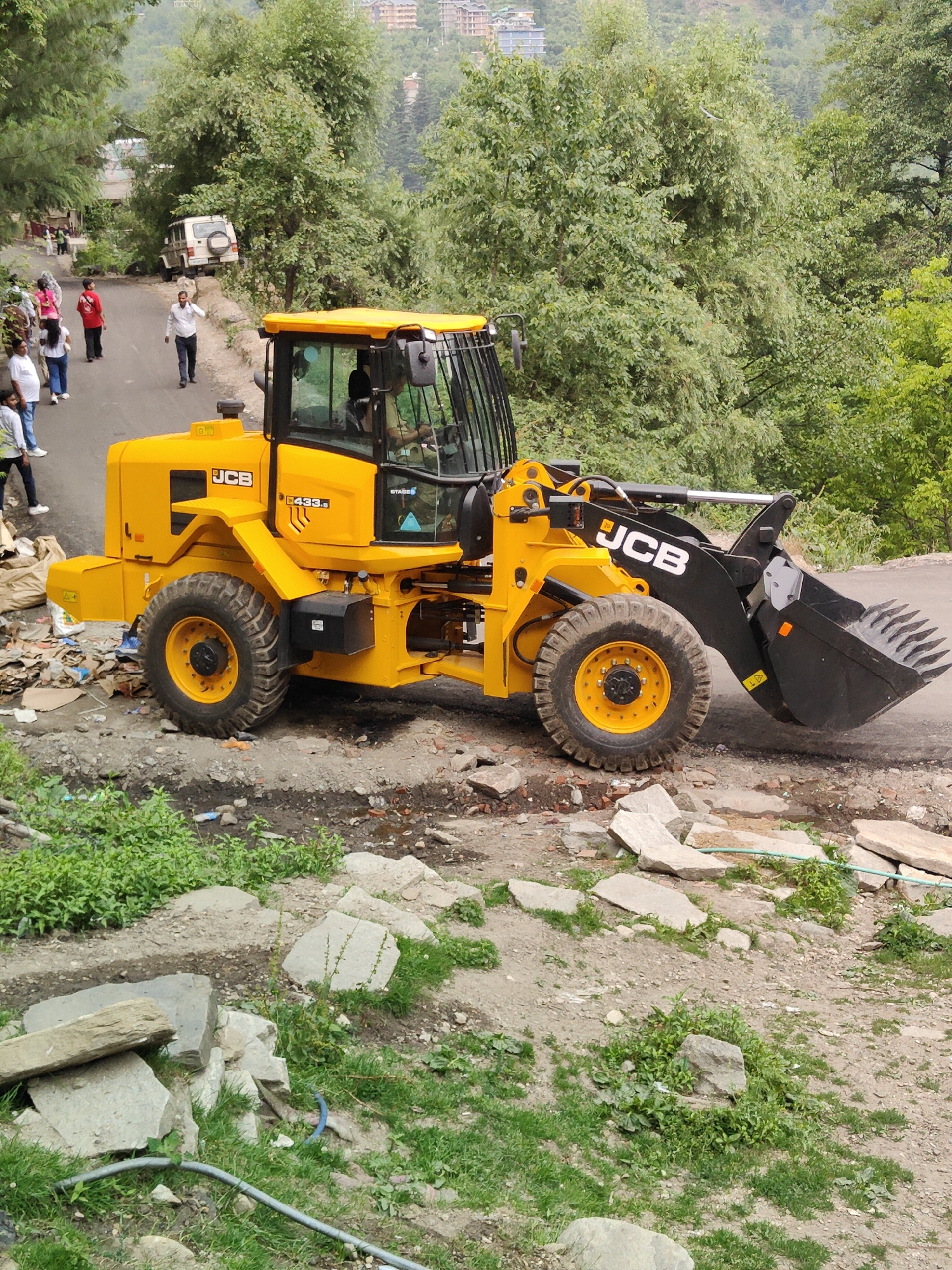 An eye-level, medium shot captures a yellow JCB 433S wheel loader parked on a paved road in Vashisht, India. The loader faces right, its front bucket extended slightly. Debris, including plastic bags and cardboard, is scattered to the left of the machine. In the background, a white SUV is parked on the side of the road, and several individuals are walking uphill on the road. Lush green trees and foliage line both sides of the road, with buildings visible on a hillside in the far distance. The ground is a mix of paved road, dirt, and scattered stones.