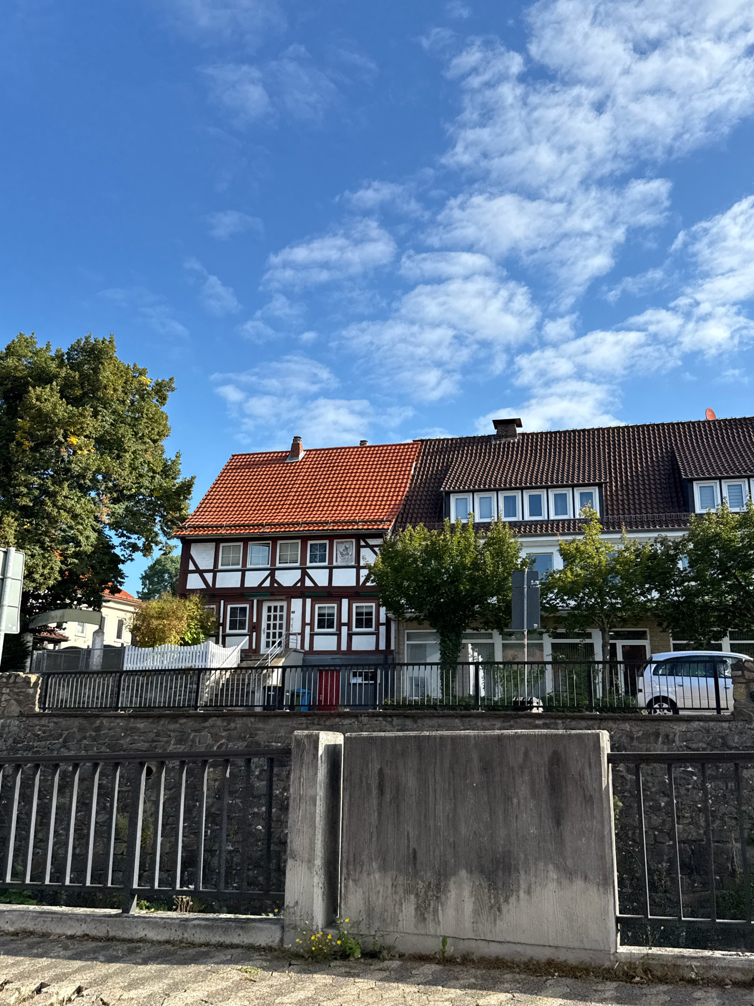 The image shows a view of a street in Osterode am Harz, Germany. On the right side of the image, there are two connected houses. The house on the left has a red tiled roof and a white facade with dark brown timber framing. It has several windows on each floor. The house on the right has a dark brown tiled roof with dormer windows and a light-colored facade. Trees are planted in front of both houses. A white van is parked in front of the right house. In the foreground, there is a stone wall with a metal fence. The sky is blue with scattered white clouds. The image appears to have been taken during the daytime.