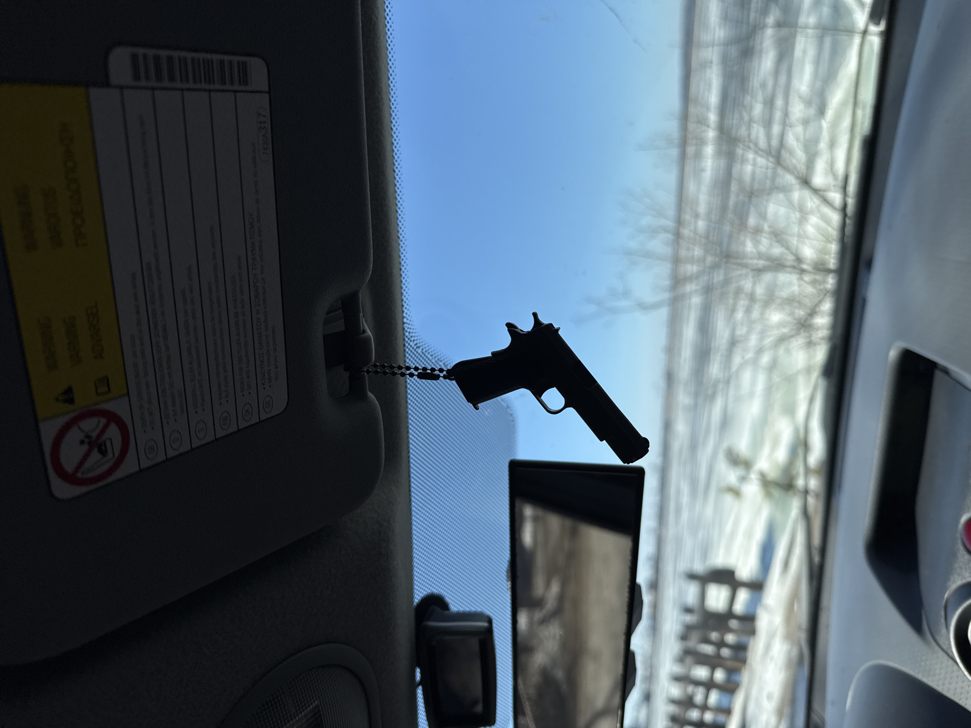 The image, taken from inside a vehicle, shows a black pistol-shaped ornament hanging from the sun visor. The visor is in a down position and features a warning label with multilingual text and safety symbols. Outside the vehicle, the view through the windshield reveals a partially snow-covered landscape, with blurry outlines of trees and a building in the background. The image suggests an outdoor environment with winter conditions. The location is Chaikovsky, Russia.