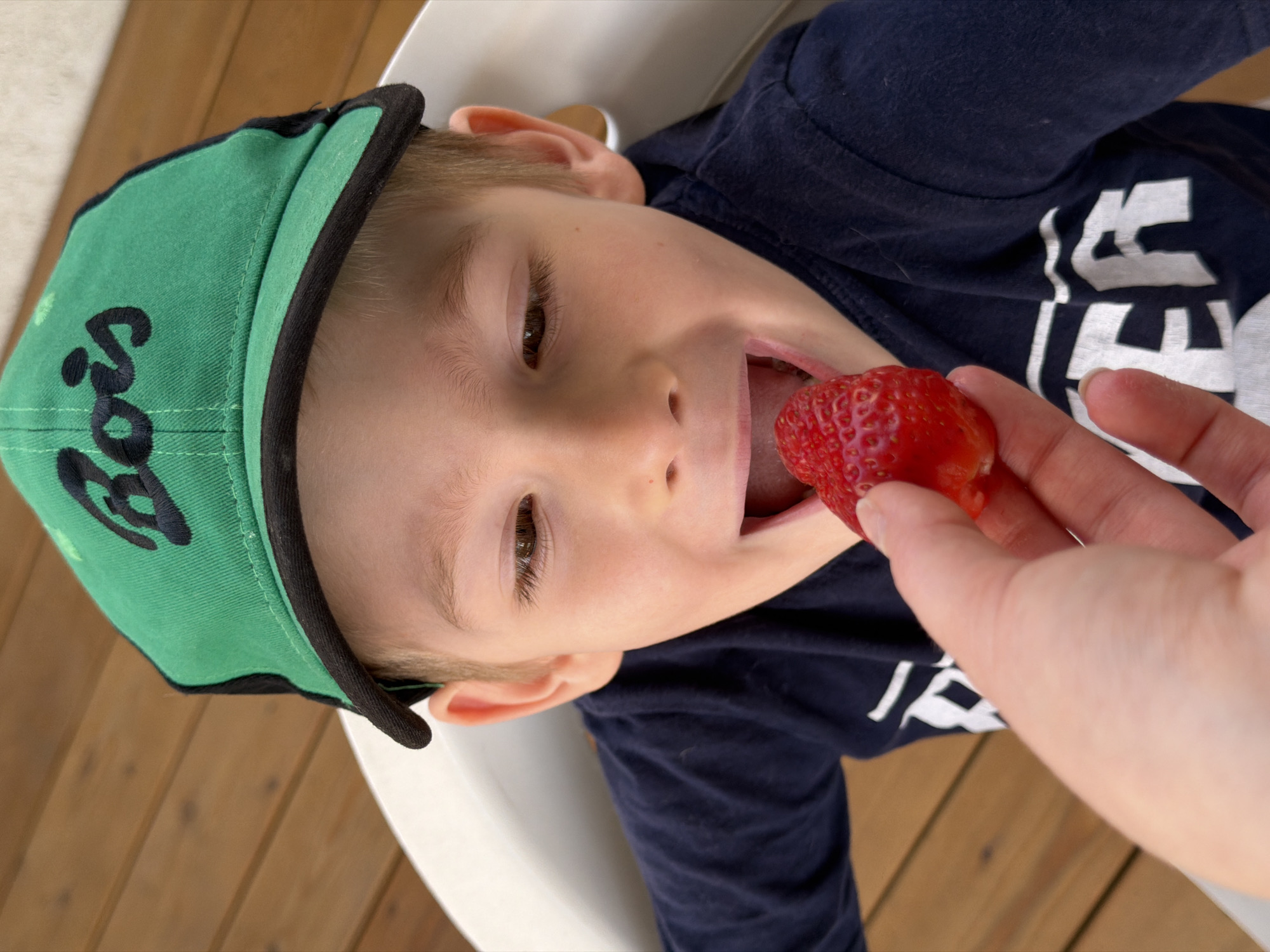 A young boy, wearing a green baseball cap with the word "Boss" embroidered on it, is being fed a strawberry. The strawberry is being held by a fair-skinned hand. The boy is wearing a navy blue t-shirt with white lettering. The location is Rossiyskiy, Russia. A wooden surface is partially visible. The boy is in a white high chair.