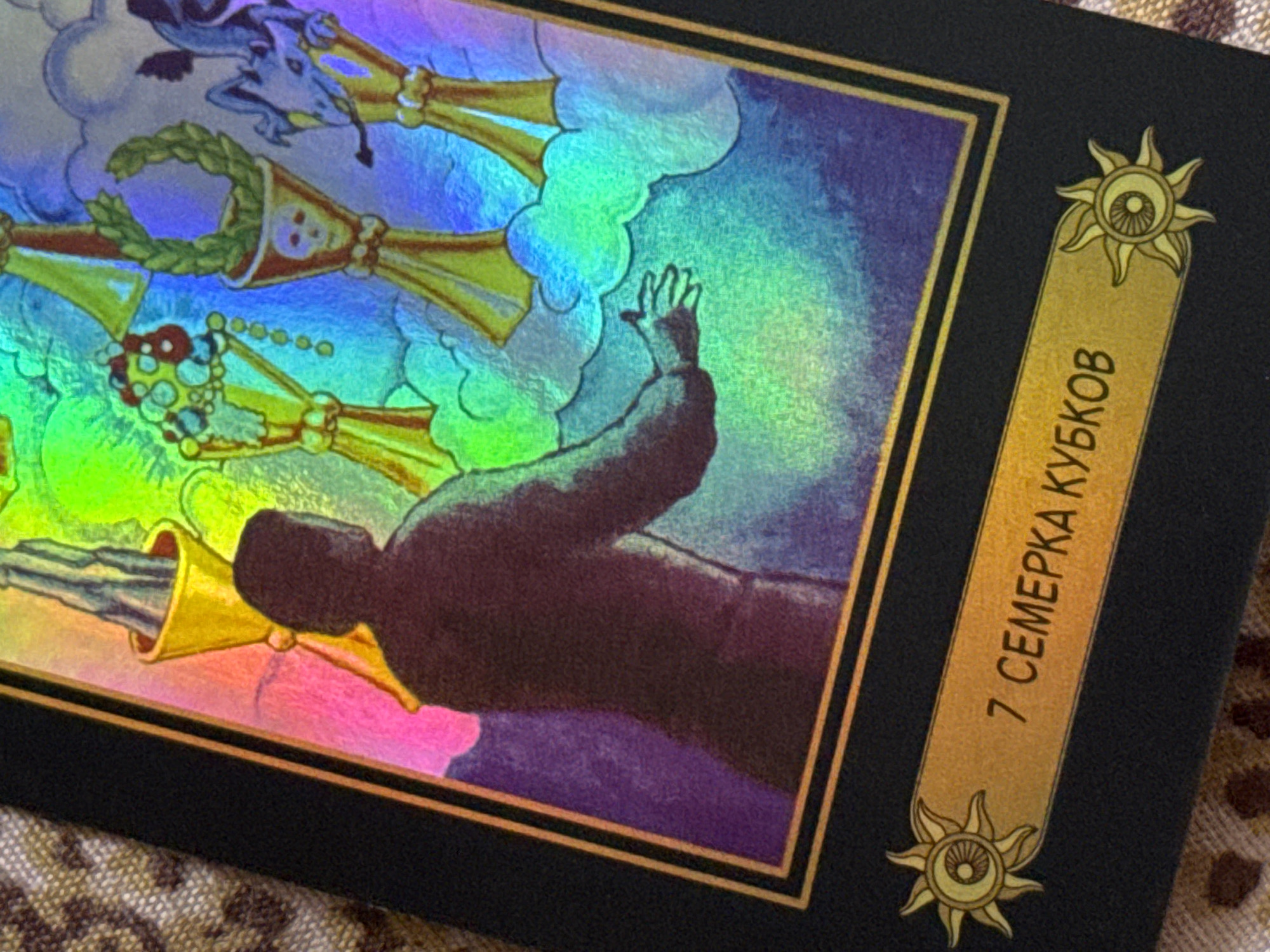 The image shows a close-up of a tarot card, likely from a Rider-Waite-Smith style deck, with a holographic or iridescent finish. The card depicts a scene with a human figure reaching upwards towards several golden trumpets adorned with wreaths and beads. The background is a swirl of colorful clouds, ranging from blues and purples to vibrant greens and yellows, suggesting a mystical or dreamlike setting. The overall impression is one of aspiration, challenge, or possibly a spiritual awakening.
On the right side of the card, a vertical gold banner displays text in Cyrillic script. The visible portion reads "7 СЕМЕРКА КУБКОВ", which translates to "7 of Cups". Below the banner is a decorative sun-like symbol with an eye in the center. The card is bordered in black with a thin gold line.
The context of Izhevsk, Russia, does not provide specific clues about the scene depicted on the tarot card itself, as the imagery is symbolic and not tied to a particular geographical location. The lighting suggests it might be daytime, but the otherworldly nature of the artwork makes it difficult to determine a specific time or weather conditions. The emotion conveyed by the human figure appears to be one of effort or longing.