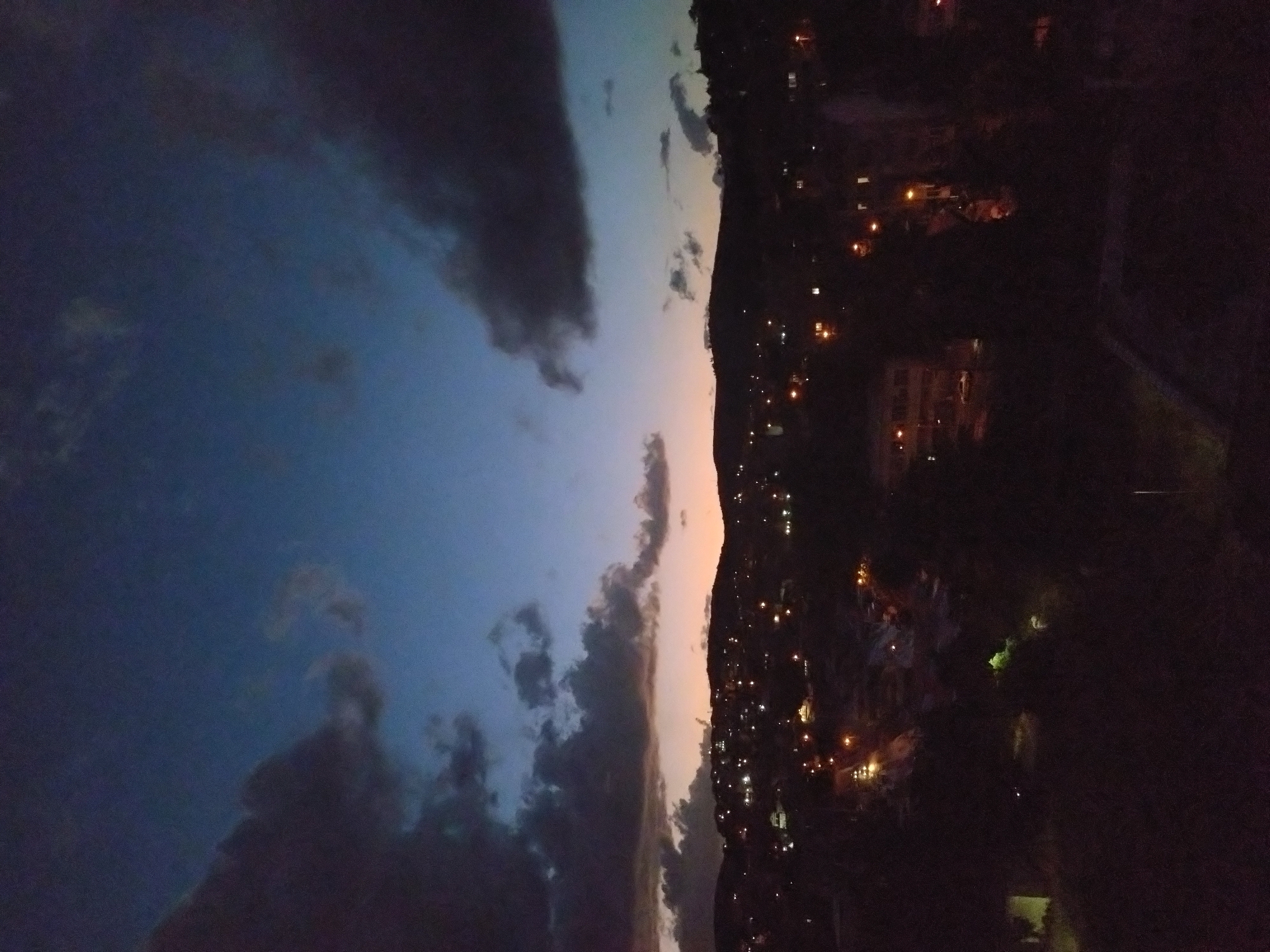 The image shows a night view of Stara Zagora, Bulgaria, with a focus on the skyline and a section of the cityscape. The sky transitions from a dark blue with heavy, shadowed clouds to a lighter, almost orange-tinted horizon near the mountainous terrain. The city's lights are scattered across the dark landscape, suggesting residential or urban areas. The lower portion of the image is very dark, obscuring the ground details.