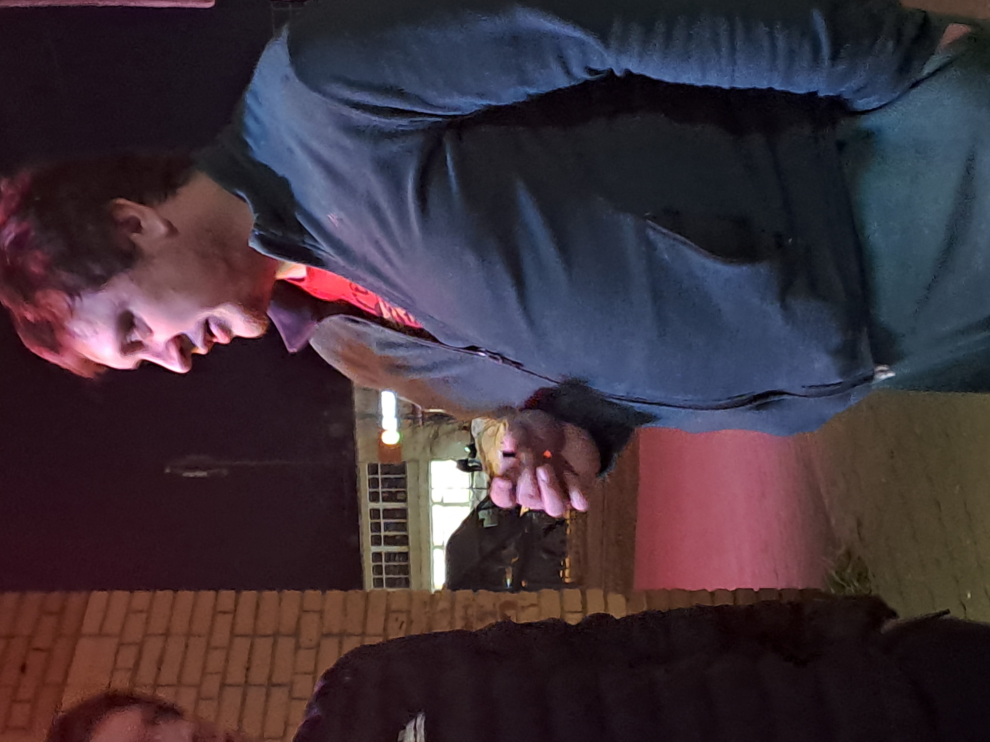 The image depicts an outdoor nighttime scene in Minino, Russia. Two male individuals are present. The primary subject, positioned on the right, is shown from the chest up in left profile. He has reddish-brown hair and light stubble, wearing a dark blue zip-up jacket over a red t-shirt. He holds a lit cigarette between the index and middle fingers of his left hand, its tip glowing orange. His mouth is slightly open. To his left, a second male is partially visible, showing his left shoulder and the side of his head, dressed in a dark, possibly padded, jacket. Behind the subjects, a light-colored brick wall extends vertically. In the background, beyond the brick wall, is a building with multiple brightly lit windows, and a dark vehicle, resembling a van, is parked in front of it. The ground surface in the immediate foreground is a reddish-purple, illuminated by artificial light.