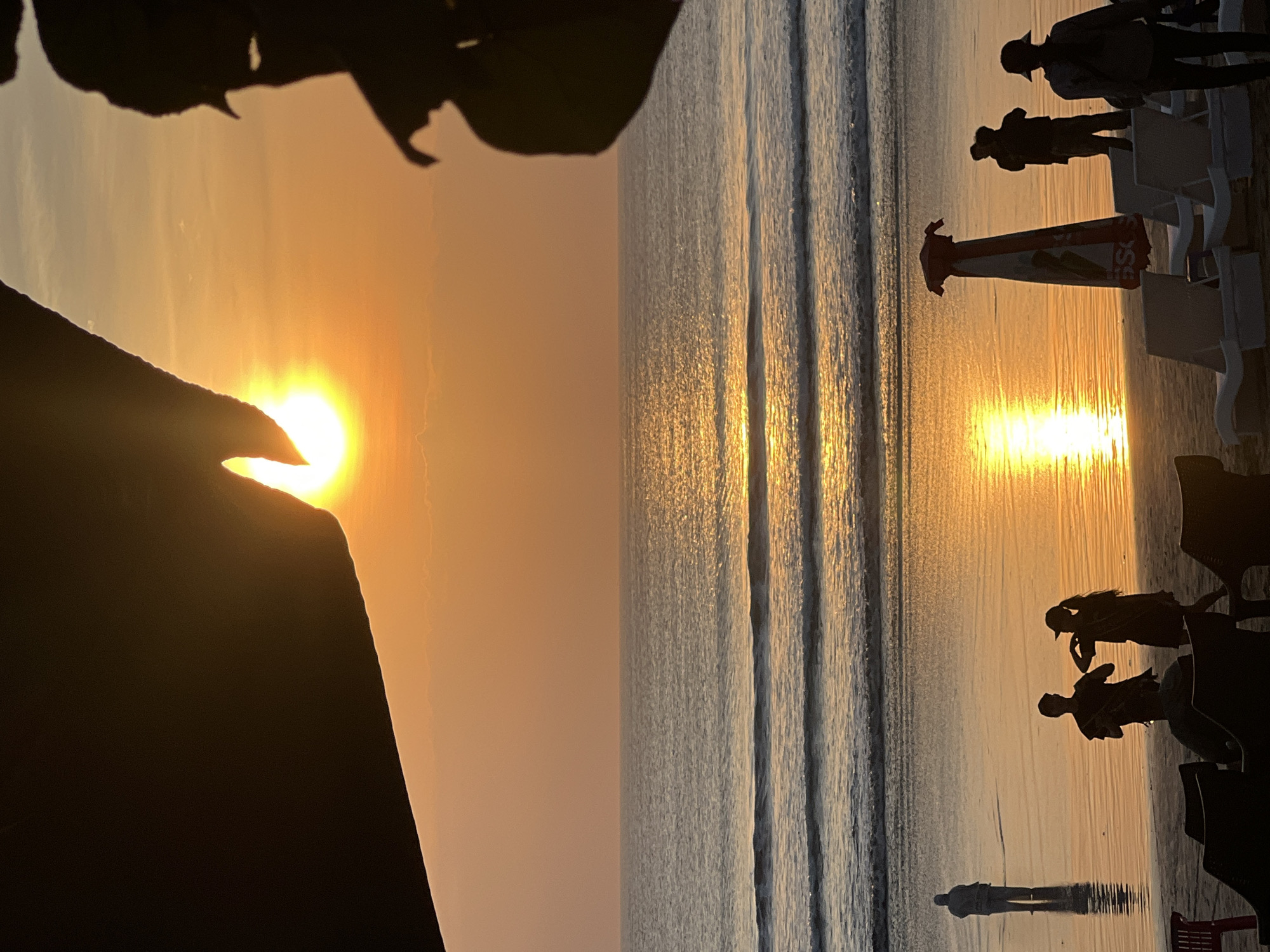 This image depicts a sunset on Kuta Beach, Indonesia. The sun is setting over the ocean, casting a warm orange glow across the sky and water. The foreground is silhouetted against the bright sky and features several people on the beach. In the middle ground, there are a few beach chairs and an umbrella. The ocean has gentle waves, and the beach is wet, reflecting the sunset. The overall scene is peaceful and serene.
