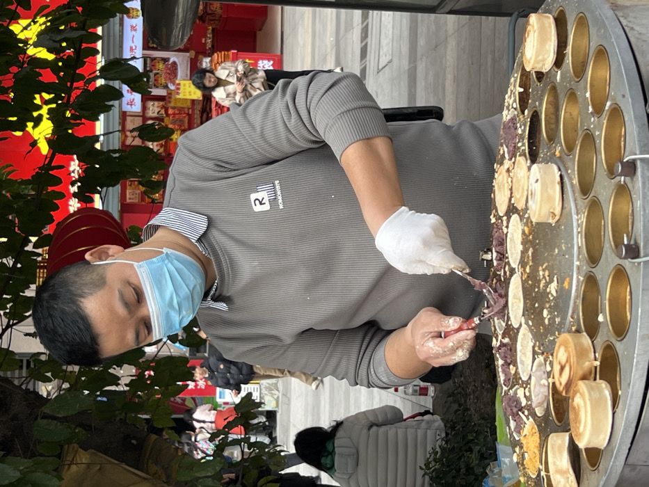 A person wearing a light blue surgical mask and white gloves is filling round molds on a large, circular griddle with a purple filling, likely for making small cakes or pastries. The person is wearing a gray long-sleeved shirt. Red decorations and a red lantern are visible in the background. A few other people are walking nearby. The street or outdoor setting is visible, suggesting a street food vendor.