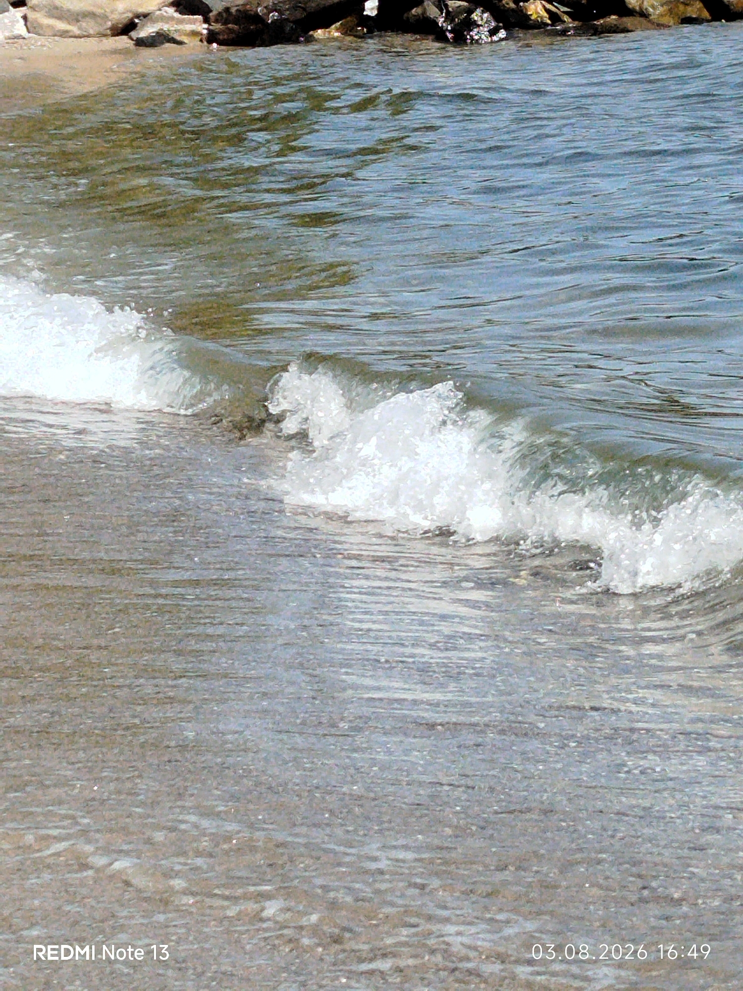 A close-up shot captures a gentle wave breaking on a sandy beach with scattered rocks in the background. The water is a muted blue-green, reflecting the clear sky. The sand is a light brown, wet near the water's edge and drier further up. The wave is frothy white as it curls and collapses onto the shore. The overall scene conveys a serene, natural environment. The image also contains text overlays: "REDMI Note 13" in the bottom left corner and "03.08.2026 16:49" in the bottom right corner. Based on the location information, this scene is likely situated on a beach within the Sithonia Municipal Unit, Greece, known for its picturesque coastlines.