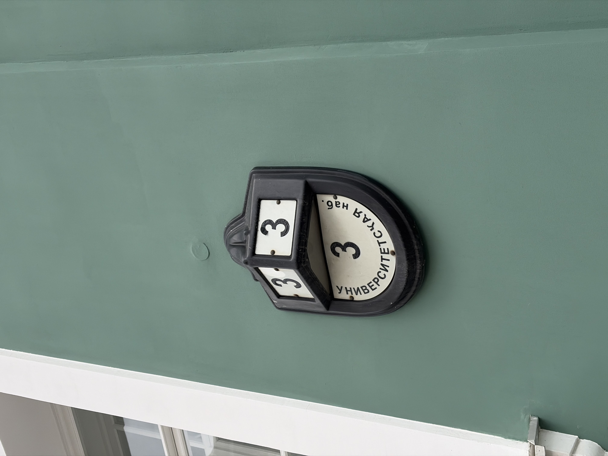 A close-up shows a black house number display attached to a green exterior wall. The display features a rotating number 3 on each of three sides of a rectangular portion. The display also has a curved, white inscription that reads "Университетская наб." (Universitetskaya nab.) in Russian. The inscription indicates that the image is likely located on University Embankment in Saint Petersburg, Russia. A section of a white window frame is visible at the bottom edge of the image.