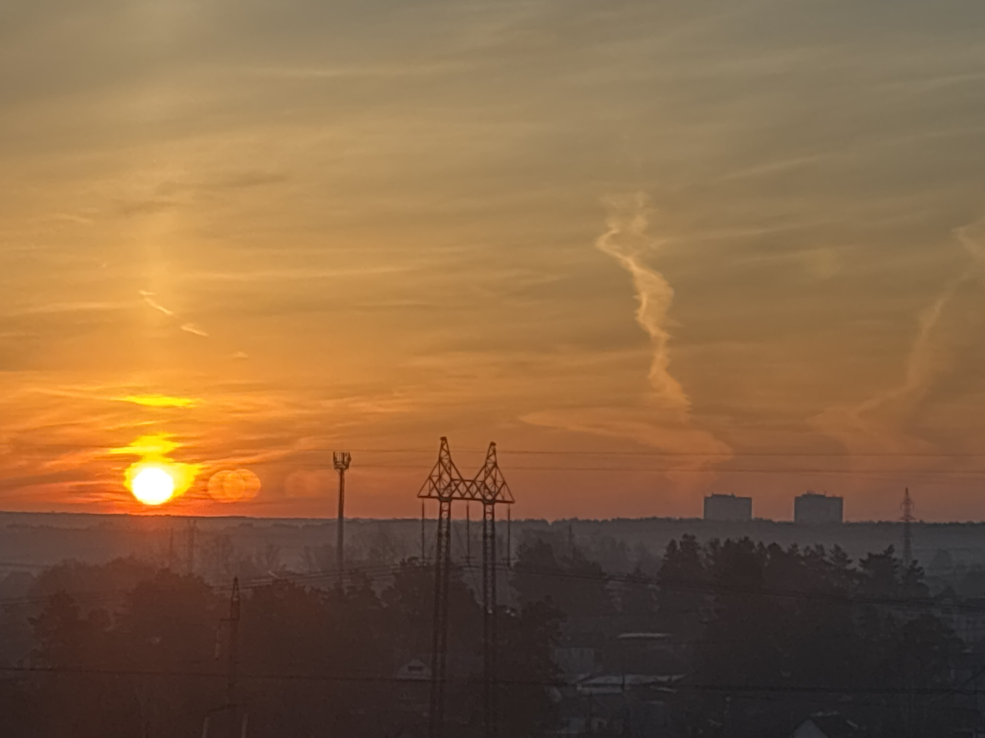 The image captures a hazy sunrise or sunset scene in what appears to be the city of Chaikovsky, Russia, identifiable by the distinct silhouette of a power plant in the distance. The sun, a bright orb, is partially obscured by a thin layer of mist or cloud, casting an orange glow across the sky. Several contrails are visible above. In the foreground, silhouetted against the light, are trees, utility poles, and electrical transmission towers. Some buildings are also visible in the lower part of the image, adding to the urban landscape. The overall scene is one of an industrial city waking up or winding down at dusk.