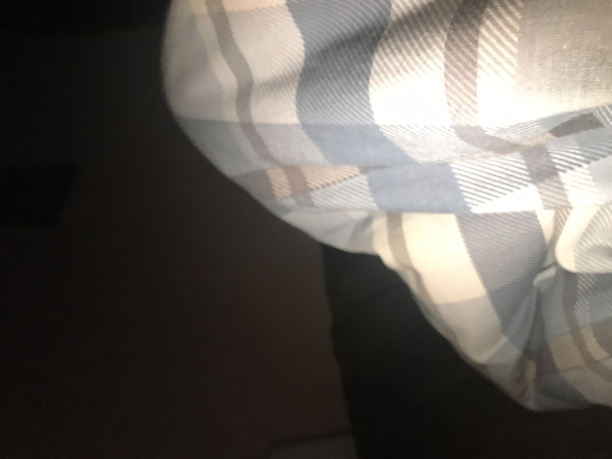 A close-up shot captures a portion of what appears to be a plaid shirt or fabric in shades of white, blue, and brown. The material is patterned with intersecting lines forming a grid-like pattern. The lighting is somewhat dim, especially on the left side of the frame, which is mostly dark. This part of the image does not allow for any information about location to be inferred. Without further visual context or identifiable landmarks, it's impossible to place the scene within the city of Yerevan, Armenia. The focus is entirely on the textile itself.
