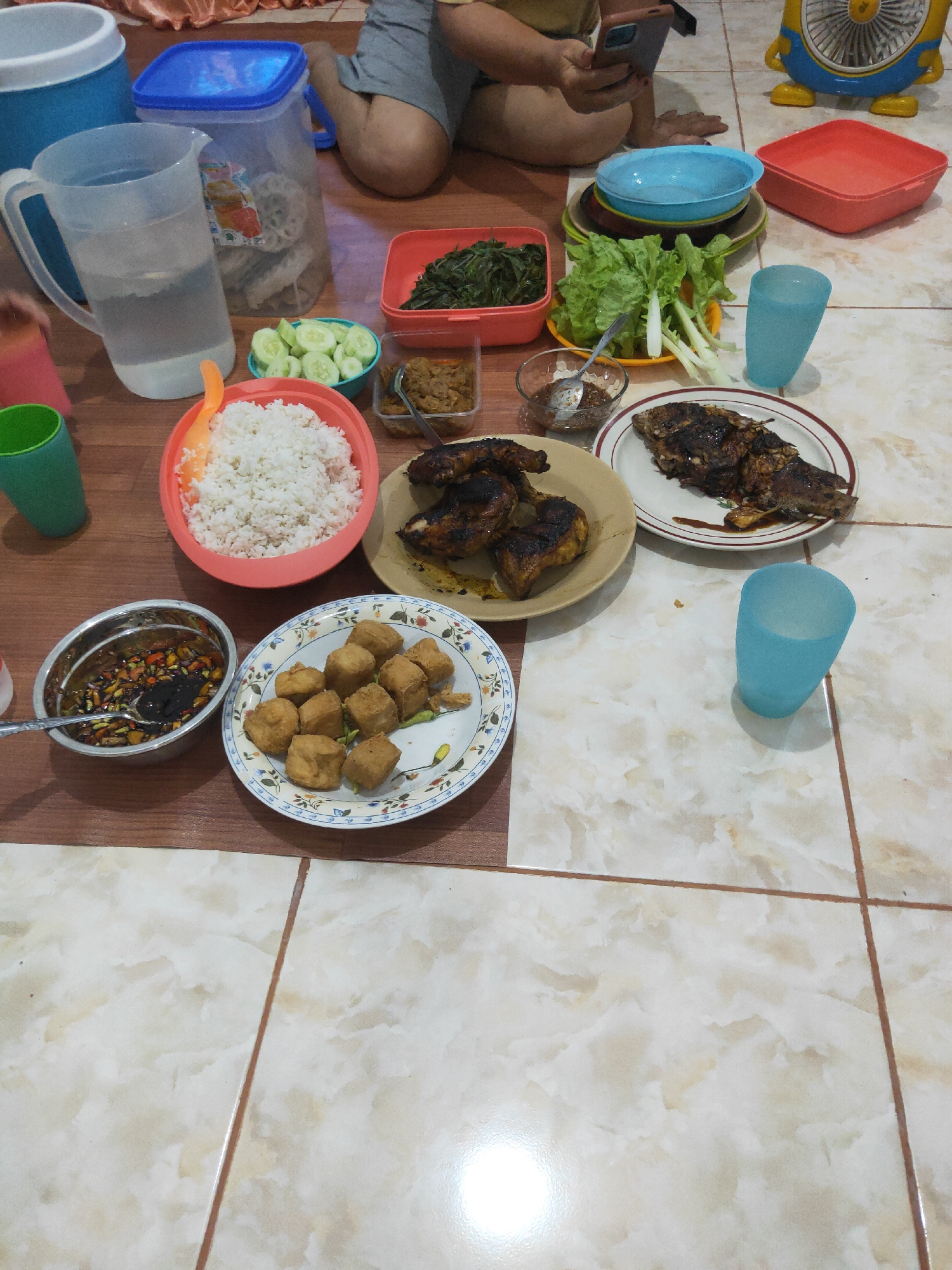 This is a high-angle, eye-level shot of a meal spread out on a tiled floor, likely in Sintang, Indonesia. The scene appears to be an informal gathering, possibly a family meal. Several dishes are visible, including a large bowl of white rice, a plate of fried tofu, grilled chicken pieces, a whole grilled fish, a bowl of vegetables (possibly water spinach), a salad of lettuce and what appear to be scallions, and a small bowl of sauce. There are also two blue plastic cups, one filled with water. To the left, a large pitcher of water sits next to a large blue storage container, and a smaller red container is to the right of the meal. In the background, a person's legs are visible, suggesting they are sitting on the floor. The tiled floor has a light brown and cream marble pattern. A small, brightly colored cartoon-like fan is visible in the upper right corner of the image. The overall impression is one of a casual, home-cooked meal shared among people sitting on the floor.
