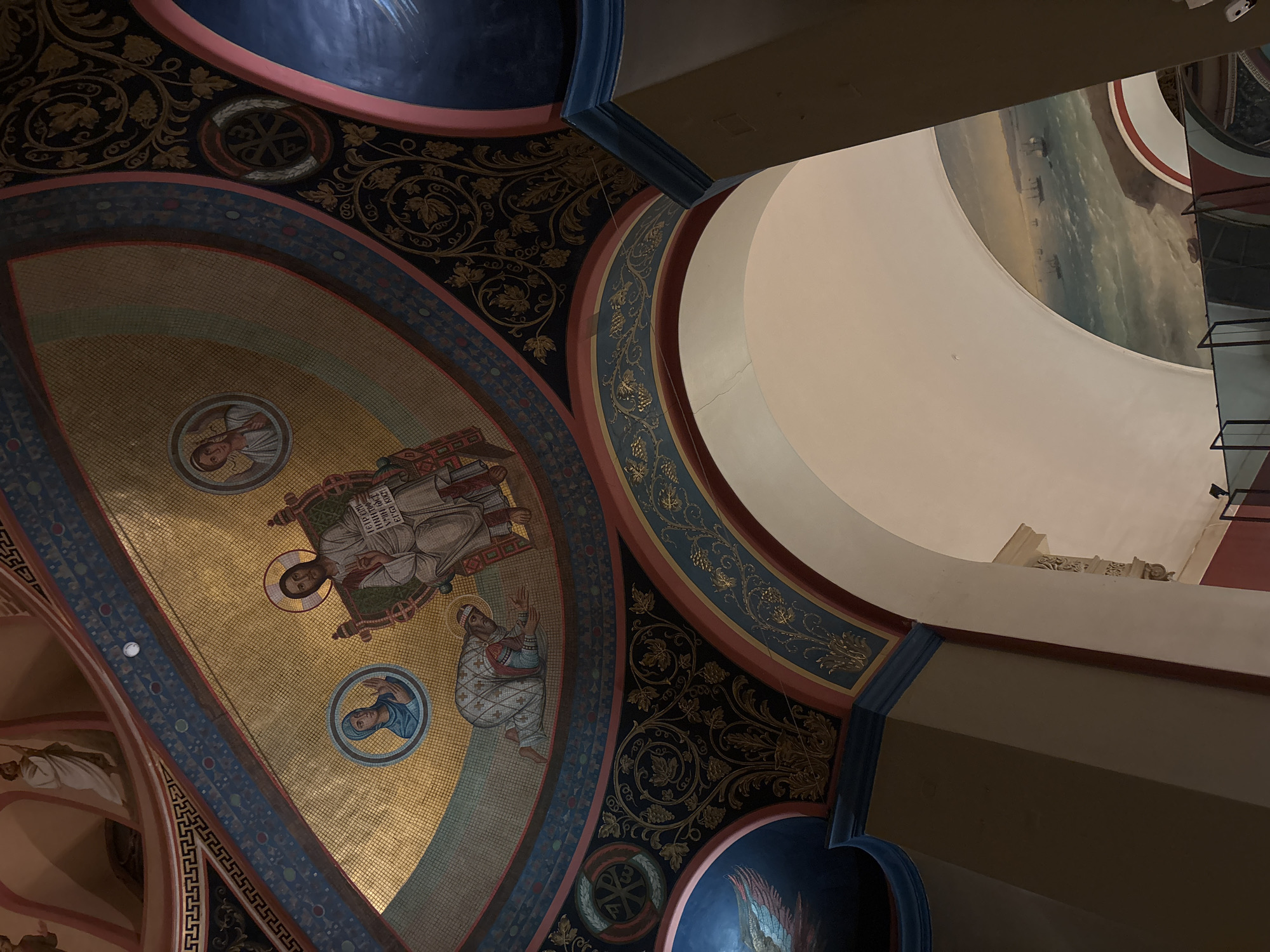 The image depicts the interior of the Cathedral of Christ the Saviour in Moscow, Russia, focusing on the ornate ceiling. The ceiling features detailed mosaics depicting religious figures. There is intricate decorative painting surrounding the mosaic panels, including floral motifs and other ornamental designs. Adjacent to the decorated ceiling is an architectural archway leading to another part of the cathedral, suggesting a complex, multi-tiered structure. The color palette is rich and includes gold, blue, and red, which are common colors in Orthodox church decoration. Artificial lighting is evident, illuminating the details of the ceiling.