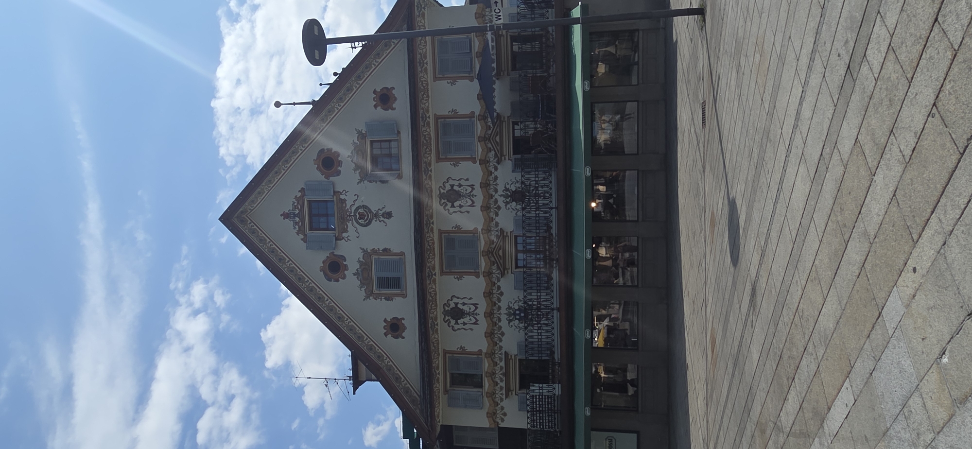 A building with traditional Bavarian-style painted decorations stands on a cobblestone plaza. The building's facade is adorned with intricate patterns in gold and red, featuring depictions of stylized floral motifs and ornate scrolls. Several windows with blue shutters are visible on the upper floors, interspersed with circular decorative elements. A balcony with a black wrought-iron railing runs along the second story, furnished with chairs and tables. Below the balcony, a green awning shades a storefront with large display windows reflecting the surroundings. A modern street lamp with a circular light fixture stands to the right of the building, casting a shadow on the plaza. A sign with "WC" and an arrow indicates a restroom facility. The sky above is a bright blue with scattered white clouds, and a sunbeam creates a lens flare on the right side of the frame. The image appears to be taken in the city of Dornbirn, Austria, given the building's architectural style and its location on a prominent plaza.