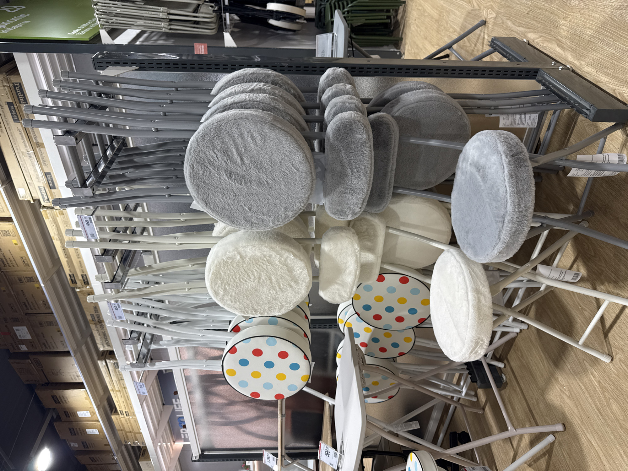 The image shows a retail store interior, likely a furniture or home goods store, in Baan Chaweng Noi, Thailand. Displayed prominently are numerous folding stools with round, padded seats. Some stools have white, fluffy seats, while others have grey, fluffy seats. Several stools feature colorful polka dot patterns on their seats, with red, yellow, and blue dots on a white background. These stools are arranged on a metal shelving unit and also placed on the floor. In the background, more folding stools and some folding tables are visible. Stacks of cardboard boxes are seen on shelves above the display, suggesting a warehouse or backstock area. The flooring appears to be a light-colored wood laminate. The lighting is artificial, consistent with an indoor retail environment. There are no people visible in the image.