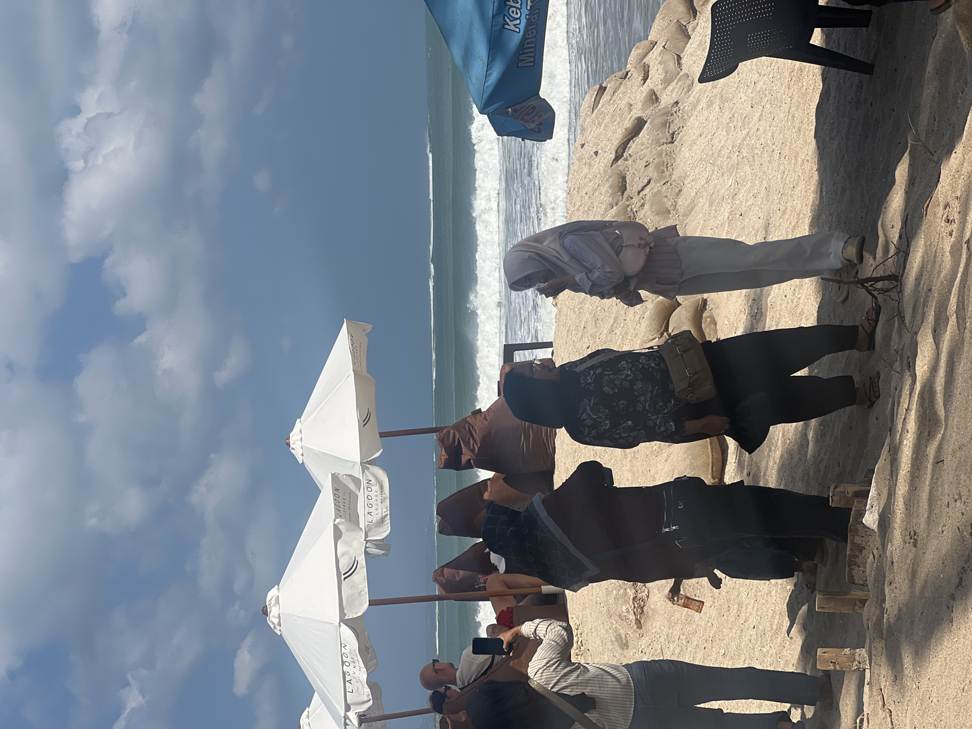 In Kuta, Indonesia, on a bright, cloudy day, a group of people are gathered on a sandy beach near the ocean. In the foreground, three women are conversing, two wearing hijabs. One woman on the left is taking a photo with her phone. In the middle ground, a man with a shaved head sits in a beanbag chair under a white umbrella. Behind him, other beanbag chairs are visible, suggesting a lounging area. The ocean in the background shows moderate waves, and several white umbrellas are set up along the shore. A blue umbrella with writing on it is visible on the right side of the image. The scene is characteristic of a beachfront cafe or lounge area in a popular tourist destination.