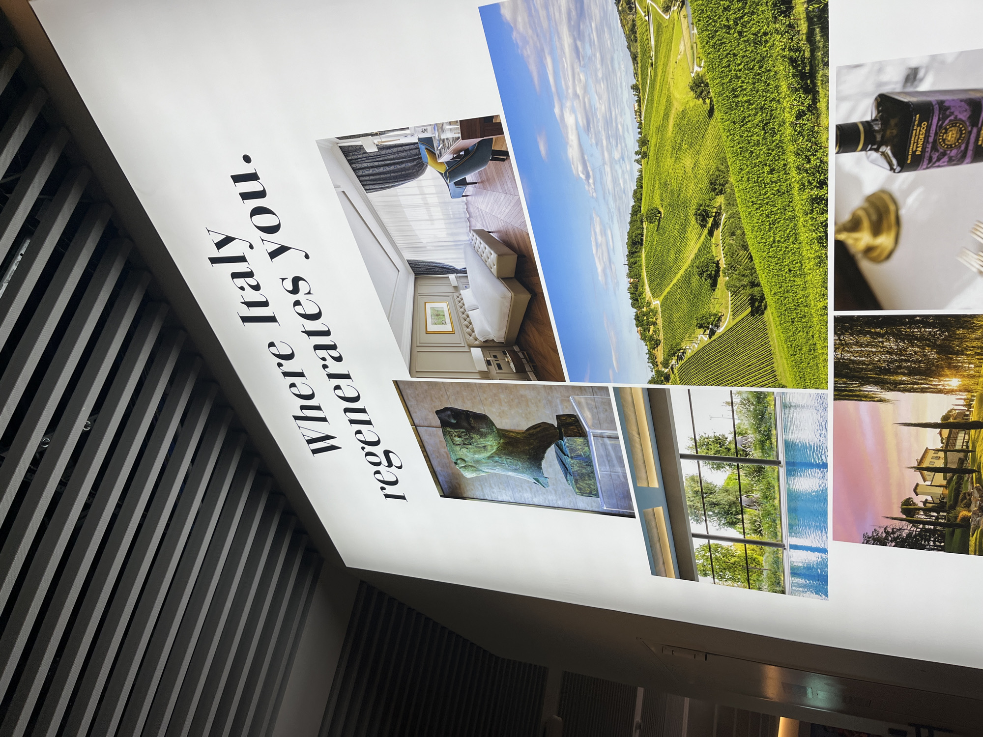A large, brightly lit promotional wall display, featuring the prominent black serif text "Where Italy regenerates you.", is observed in a modern indoor environment located in Bologna, Italy. The display is mounted on a white wall, beneath a slatted grey ceiling. Six distinct photographic panels are arranged beneath the text. These panels include, from top-left to bottom-right: a bronze bust of a classical head, a luxurious hotel bedroom with a large white bed and elegant furnishings, expansive green vineyards under a partly cloudy blue sky, an indoor swimming pool with large windows overlooking trees, a Tuscan-style villa with cypress trees at sunset, and a close-up of a dark bottle of "OLIO EXTRAVERGINE DI OLIVA PANDESCO" on a white tablecloth alongside dining items. No individuals are present in the scene.
