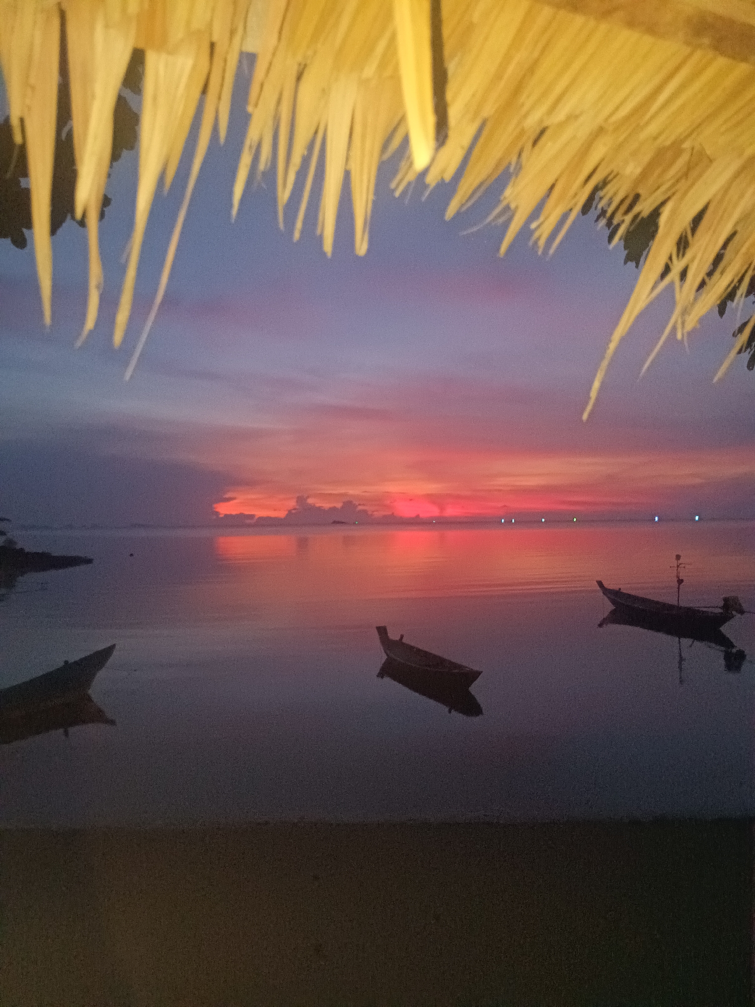 The image captures a serene sunset scene in Baan Wok Tum, Thailand. The foreground is dark, revealing a sandy beach. Above this, calm water reflects the vibrant colors of the sunset. Three traditional Thai longtail boats are visible, gently floating on the water. In the background, the setting sun paints the sky with hues of orange, red, and purple. Lights dot the horizon, likely from buildings along the coast. A thatched roof overhangs the scene, partially obscuring the sky at the top of the image.
