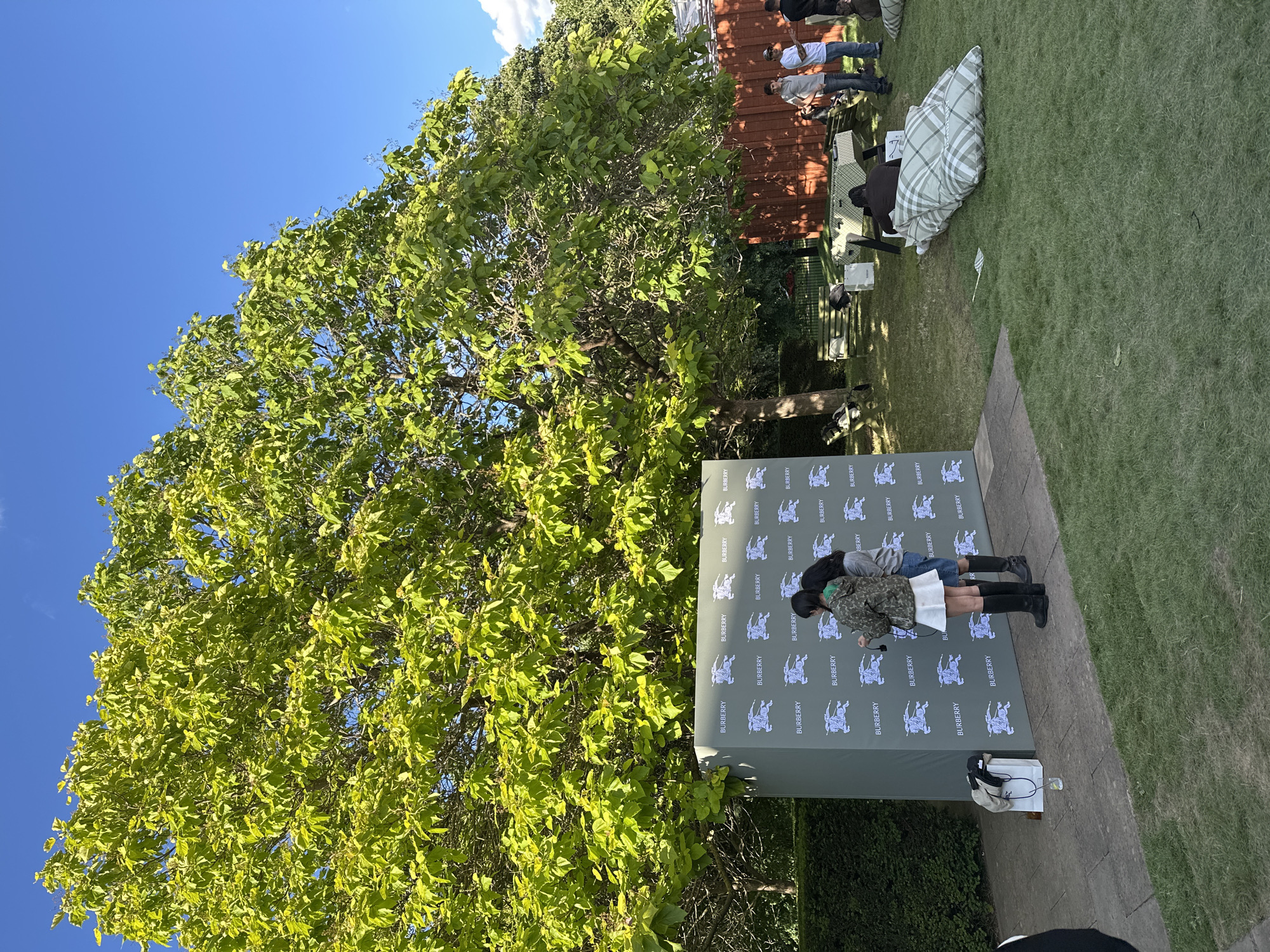 In an outdoor setting on a sunny day with a clear blue sky, a large tree with vibrant green foliage dominates the background. In front of the tree, a rectangular green backdrop is set up, branded with repeated Burberry logos and equestrian motifs. Two individuals stand near the backdrop, one in a camouflage jacket and white skirt, the other in a grey top and denim shorts, both wearing black boots. To the right of the backdrop, a group of people are seated and standing, some on large, checkered floor cushions and others near picnic tables. The ground is covered in manicured grass. The scene appears to be an event or gathering, possibly in a park or garden within London, United Kingdom.