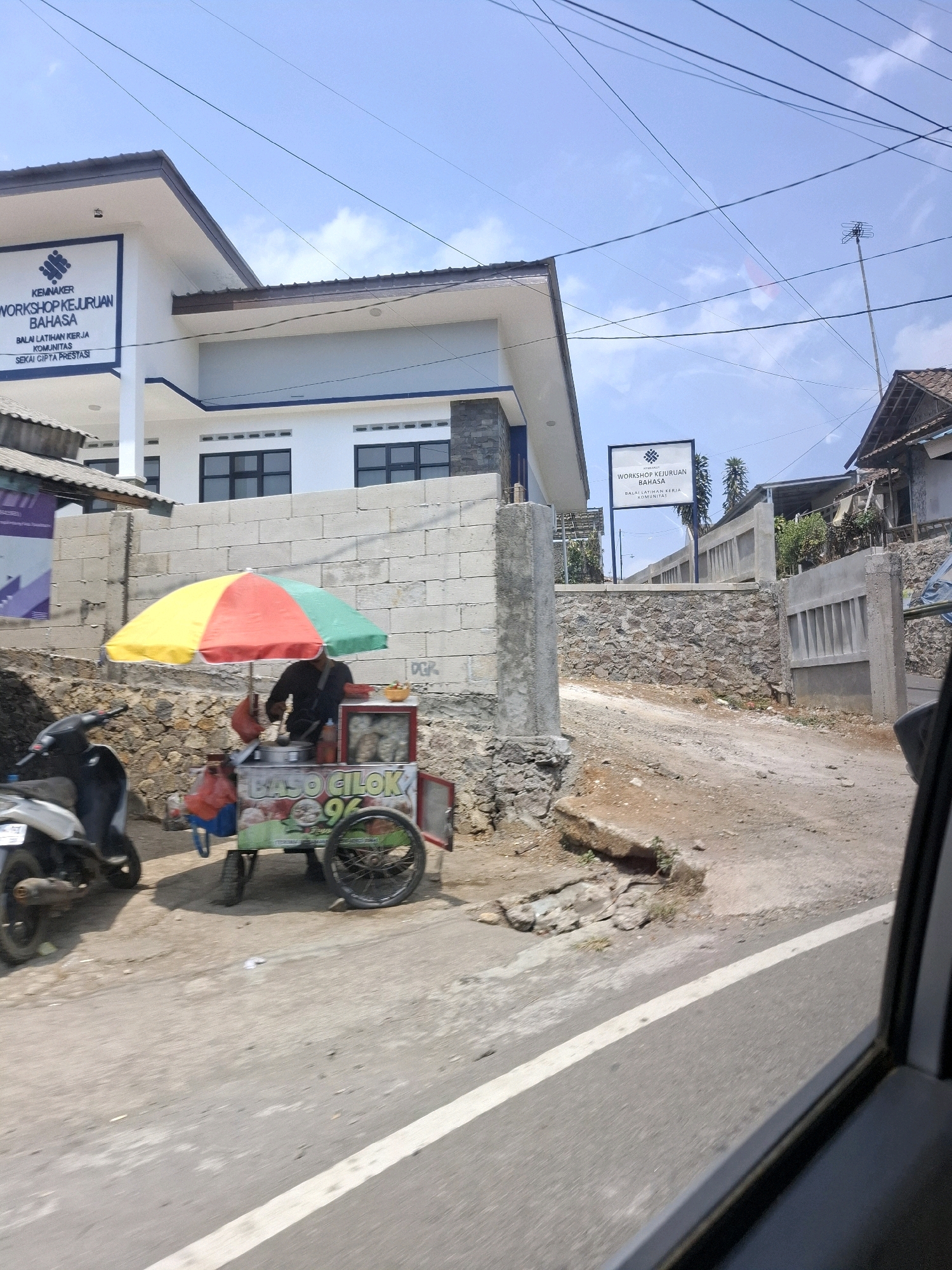 In Cijangkar, Indonesia, a street-level view captures a sunny day with a blue sky dotted with white clouds. In the foreground, a street vendor operates a mobile cart selling "BASO CILOK". The cart is adorned with a colorful umbrella and displays the price "96". A person, dressed in dark clothing, is seen preparing food behind the cart. To the left, a motorcycle is parked on the side of the road. Behind the cart and motorcycle, a rough stone wall forms a boundary. Further in the background, a modern white building with blue accents is visible, featuring a sign that reads "KEMNAKER WORKSHOP KEJURUAN BAHASA BALAI LATIHAN KERJA KOMUNITAS SEKAI CIPTA PRESTASI". The building appears to be a vocational training center. A dirt road or path leads up from the foreground towards the building, with more stone walls and some greenery visible. Overhead, a network of power lines crisscrosses the sky. The overall scene depicts a typical roadside environment in a semi-urban or rural Indonesian setting, with a blend of commercial activity and institutional buildings.