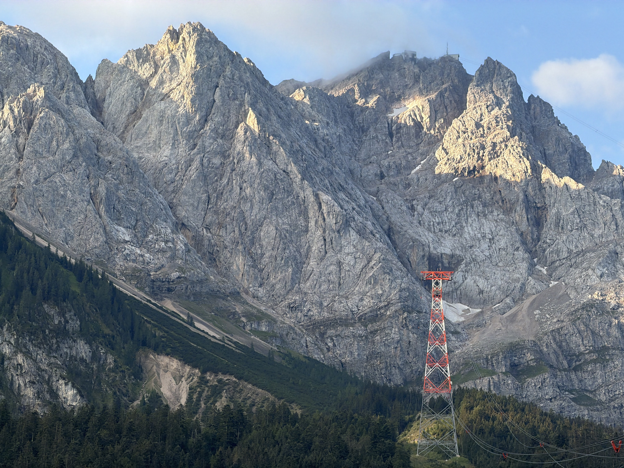 The image depicts a dramatic mountain landscape in Grainau, Germany. In the foreground, a tall, red and white lattice tower, characteristic of a cable car support structure, stands prominently. The tower is surrounded by a dense forest of dark green coniferous trees. Behind the forest, the imposing, rugged slopes of a large mountain range dominate the scene. The mountains are rocky and steep, with visible crevices and ledges. The upper portions of the mountains are illuminated by warm sunlight, casting shadows and highlighting the textures of the rock. Some patches of snow or ice are visible on the higher slopes. In the distance, at the very top of one of the peaks, a structure resembling a building or a viewing platform can be seen, partially obscured by a wispy cloud. The sky above is a clear blue with a few scattered white clouds. The overall impression is one of natural grandeur and human engineering integrated into the mountainous terrain.