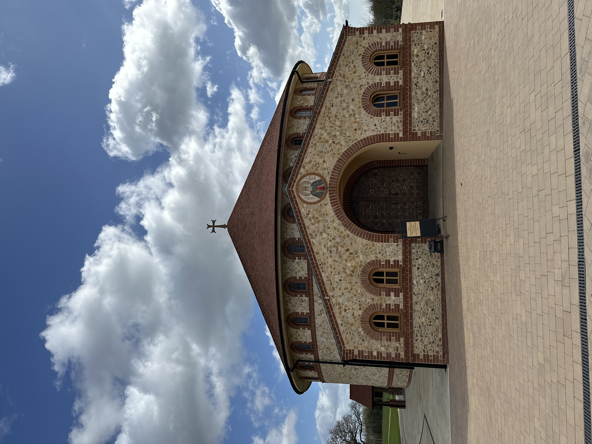 The image presents the Orthodox Monastery of St. John the Baptist in Maldon, United Kingdom, viewed under a partially cloudy blue sky. The central structure is a circular building topped with a conical, terracotta-tiled roof crowned by a single metal cross. Its facade is constructed from light-colored, irregular stone masonry interspersed with red brickwork, which forms arches, window surrounds, and horizontal banding. A prominent arched main entrance in the center features large, dark, ornate wooden double doors. Above this arch, a mosaic icon depicts two robed figures. Multiple arched windows, fitted with light-colored frames, are visible across the building's exterior.
The foreground consists of an expansive paved forecourt, composed of light brown rectangular pavers, with a dark linear drainage grate running across the bottom of the frame. To the right of the main entrance, a black A-frame sign and a small black metal rack are positioned on the paving. A black downpipe is visible along the left side of the main circular structure. To the left of the primary building, a smaller, single-story extension with a tiled roof is partially visible, adjacent to a wide expanse of green grass and distant trees. No individuals are visible in the scene.