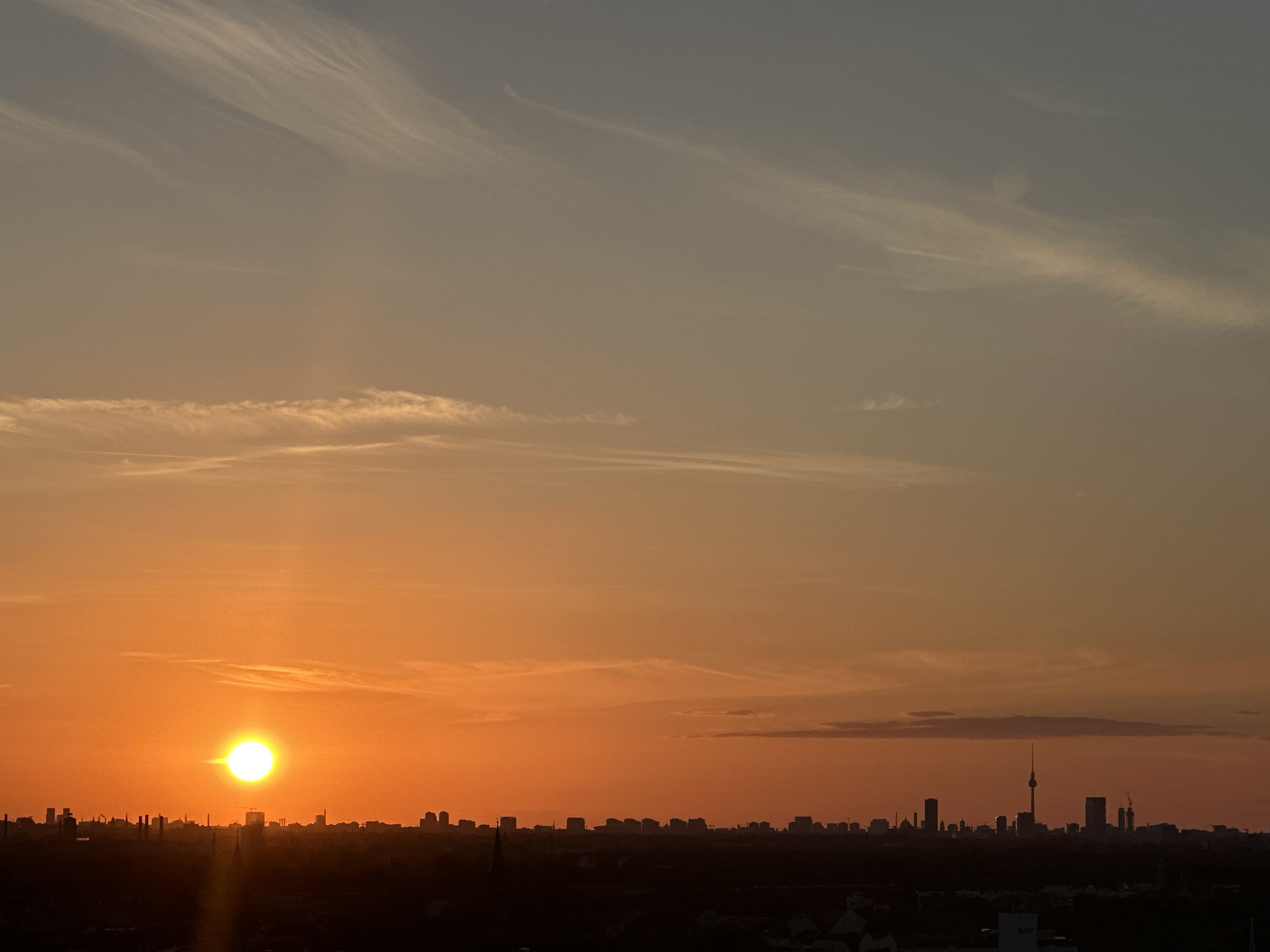 This is a long-shot photograph of a sunset over the city of Berlin, Germany. The sun, a bright orb, is low on the horizon, casting a warm orange glow across the sky. Wispy clouds are scattered across the sky, adding texture and depth to the scene. The city skyline is silhouetted against the sunset, with a prominent television tower rising above the other buildings. The overall impression is one of a tranquil evening in the city.