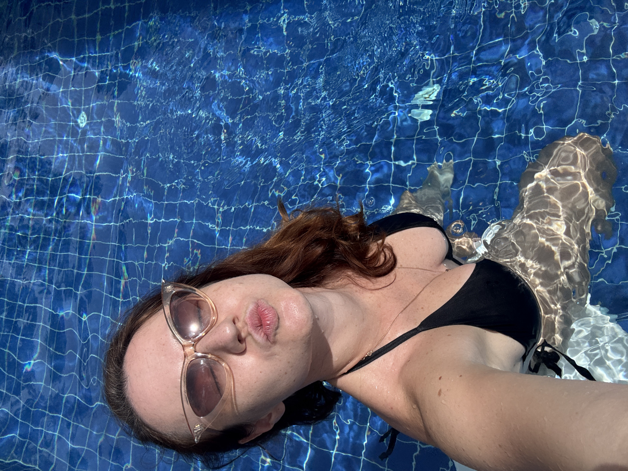 A woman in a black bikini and large cat-eye sunglasses is in a swimming pool, puckering her lips as if for a kiss. She has reddish-brown hair and is looking towards the camera. The pool water is a vibrant blue, with sunlight reflecting off the surface and creating a checkered pattern from the pool tiles. The woman is partially submerged, with water droplets visible on her skin and hair.