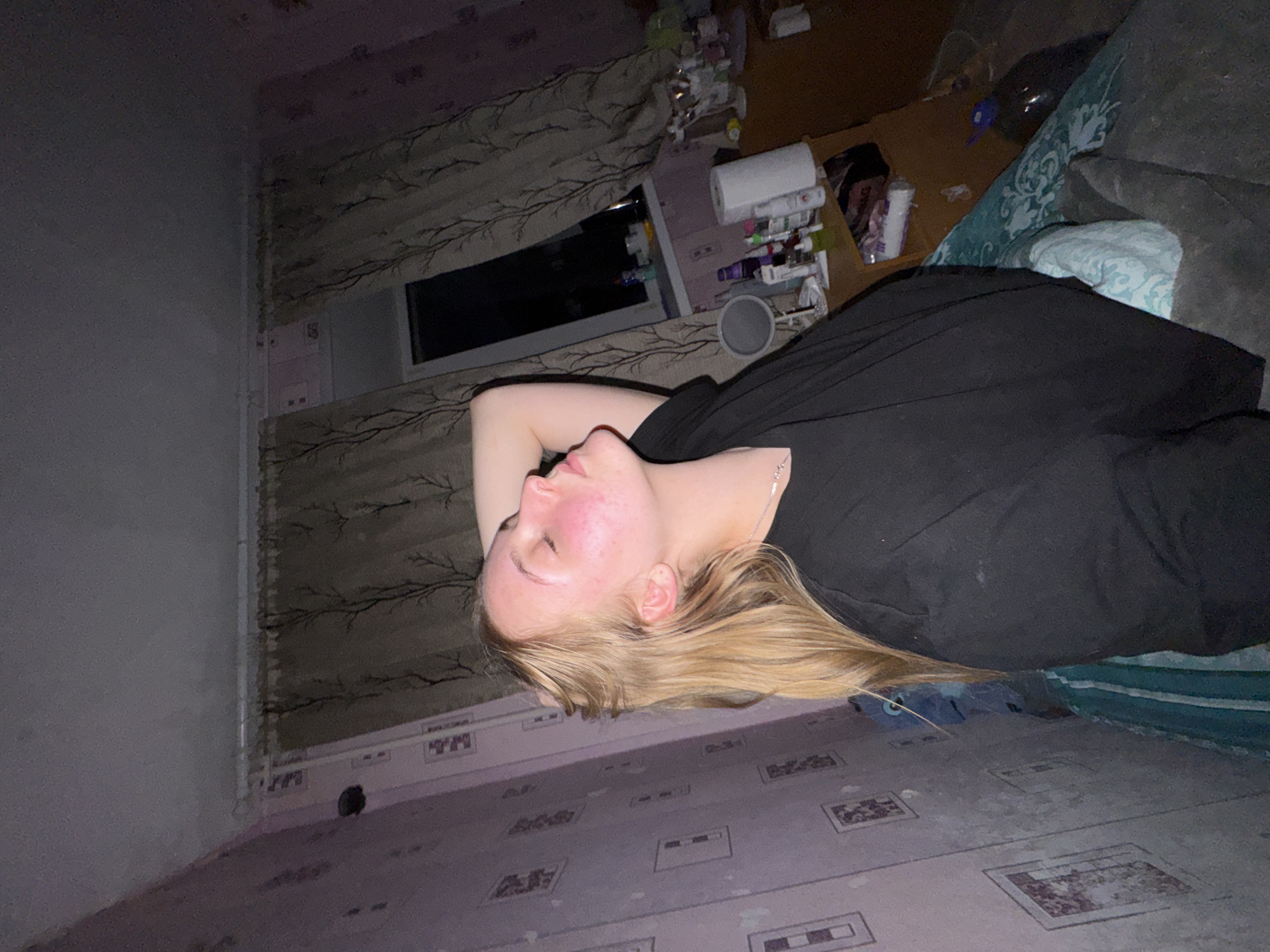 This indoor photograph, taken in Perm, Russia, depicts a young woman resting on her side in what appears to be a bedroom. She has long, light blonde hair and is wearing a black top with a thin silver necklace. Her profile shows a face with some redness, and her eyes are closed or looking downwards. One arm is bent, with her hand positioned near her head.
The woman is lying on a bed or couch, partially visible beneath her, featuring patterned bedding in shades of blue, white, and green. Beside her is a cluttered bedside table or shelf, covered with numerous bottles, containers, a white roll, and a grey cup, suggesting an accumulation of personal care products or miscellaneous items. Behind this, a rectangular window with dark panes suggests it is nighttime or a dimly lit interior, framed by curtains adorned with a repeating pattern of dark tree branches.
The room's walls vary; one is a plain, solid grey or beige, while another displays a repeating, faded square or rectangular pattern. The floor also exhibits a similar patterned surface, possibly linoleum. The overall lighting in the room is dim, creating a subdued and personal atmosphere. The scene captures a moment of rest or relaxation within a lived-in space. No discernible text is visible on any objects or surfaces in the image.