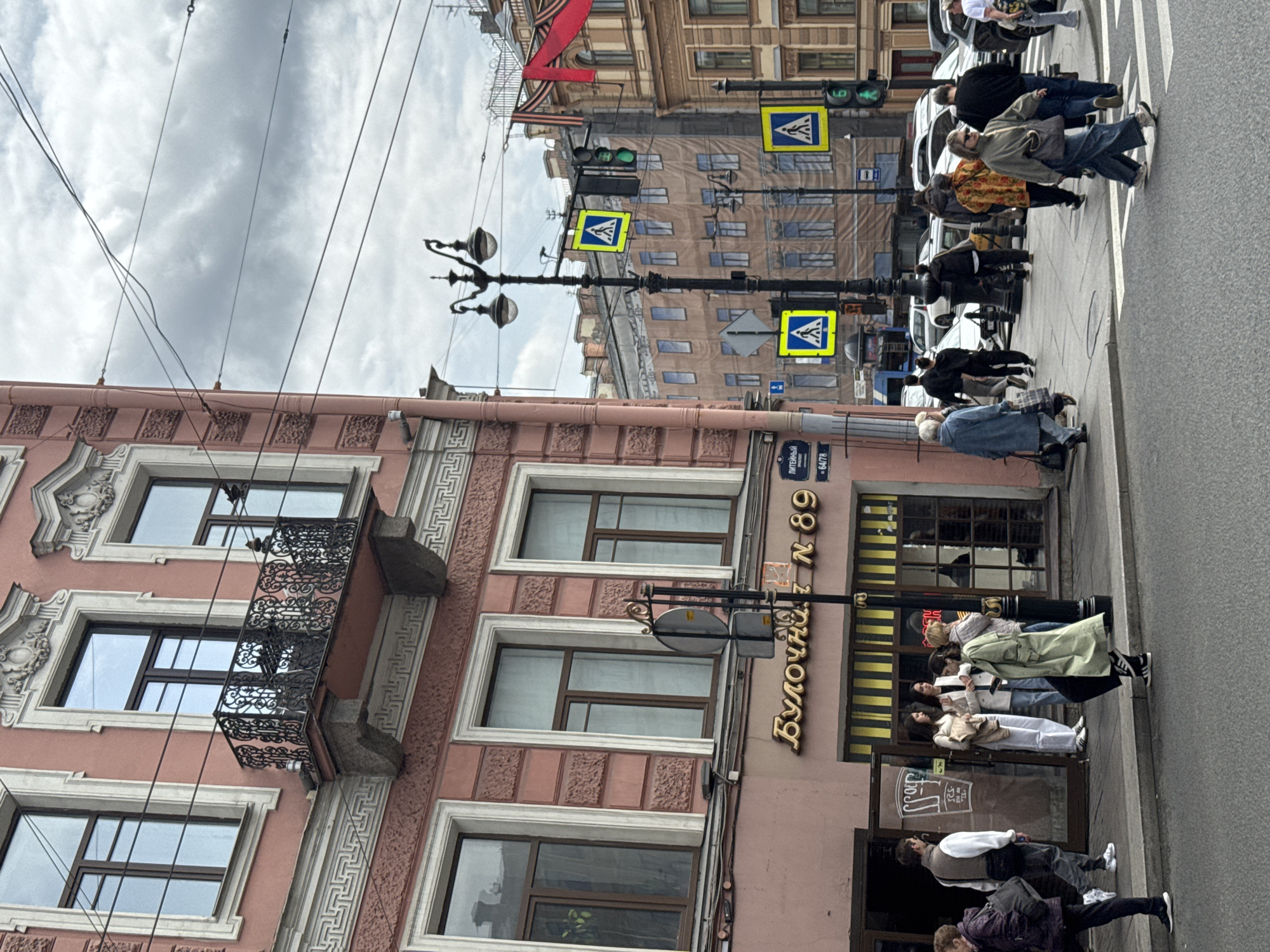 The image depicts a street scene in Saint Petersburg, Russia, with a building on the left side featuring a bakery called "Булочная N 89" (Bulochka N 89). The building has multiple windows and a decorative balcony. On the right side of the image is a street with pedestrians crossing a crosswalk. There are pedestrian crossing signs and traffic lights. The sky is overcast.