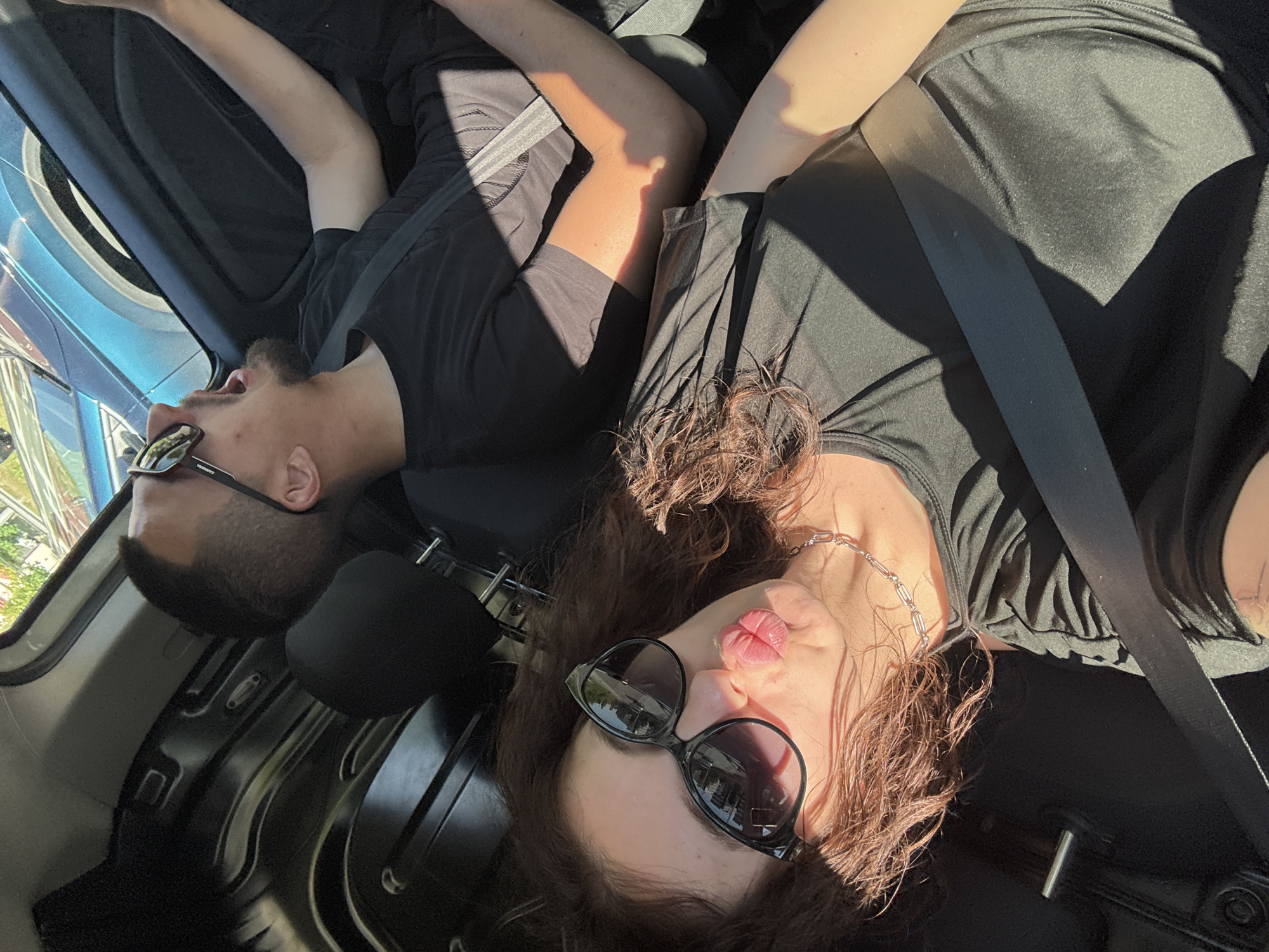 Two people are inside a vehicle. The person on the left wears a black t-shirt and sunglasses. The person on the right wears a dark-colored shirt, a silver chain necklace, and sunglasses. Both individuals appear to be reclining in the seats of the vehicle. The location and city are unknown.
