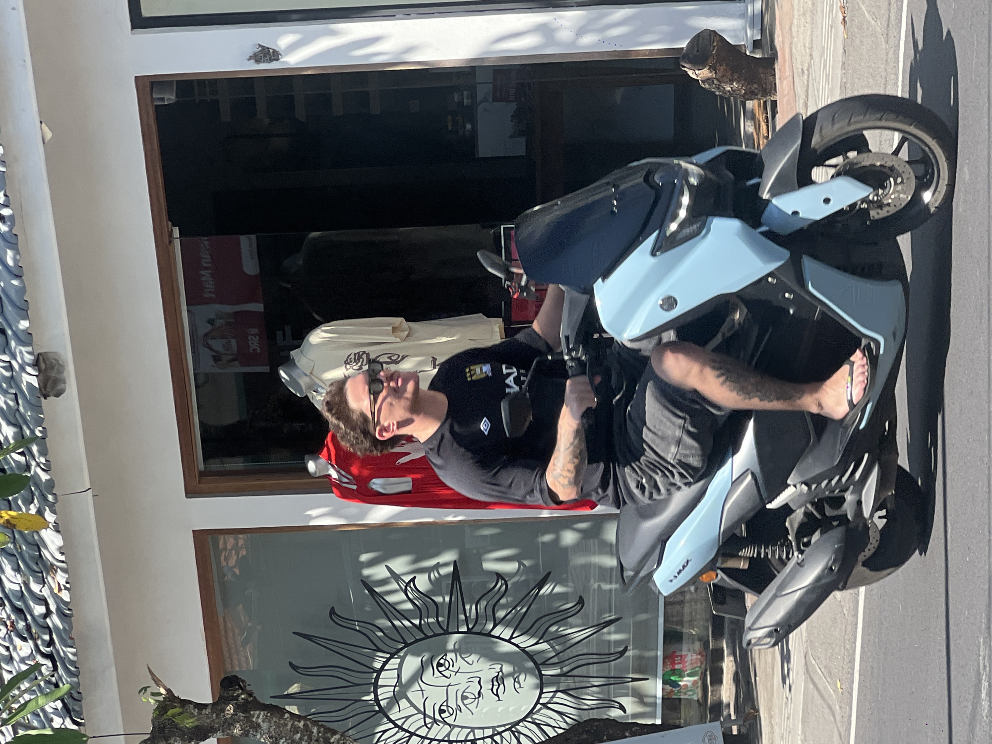 A fair-skinned man sits on a blue Yamaha TMAX scooter parked on a street in North Kuta, Indonesia. He wears a black t-shirt, dark shorts, and flip-flops, with tattoos visible on his left arm and right leg. He looks to his left. The scooter is parked in front of a store, with a window displaying a stylized sun design. A mannequin wearing a red garment and a white t-shirt is visible inside the store. The street appears paved. The overall lighting indicates a sunny day.