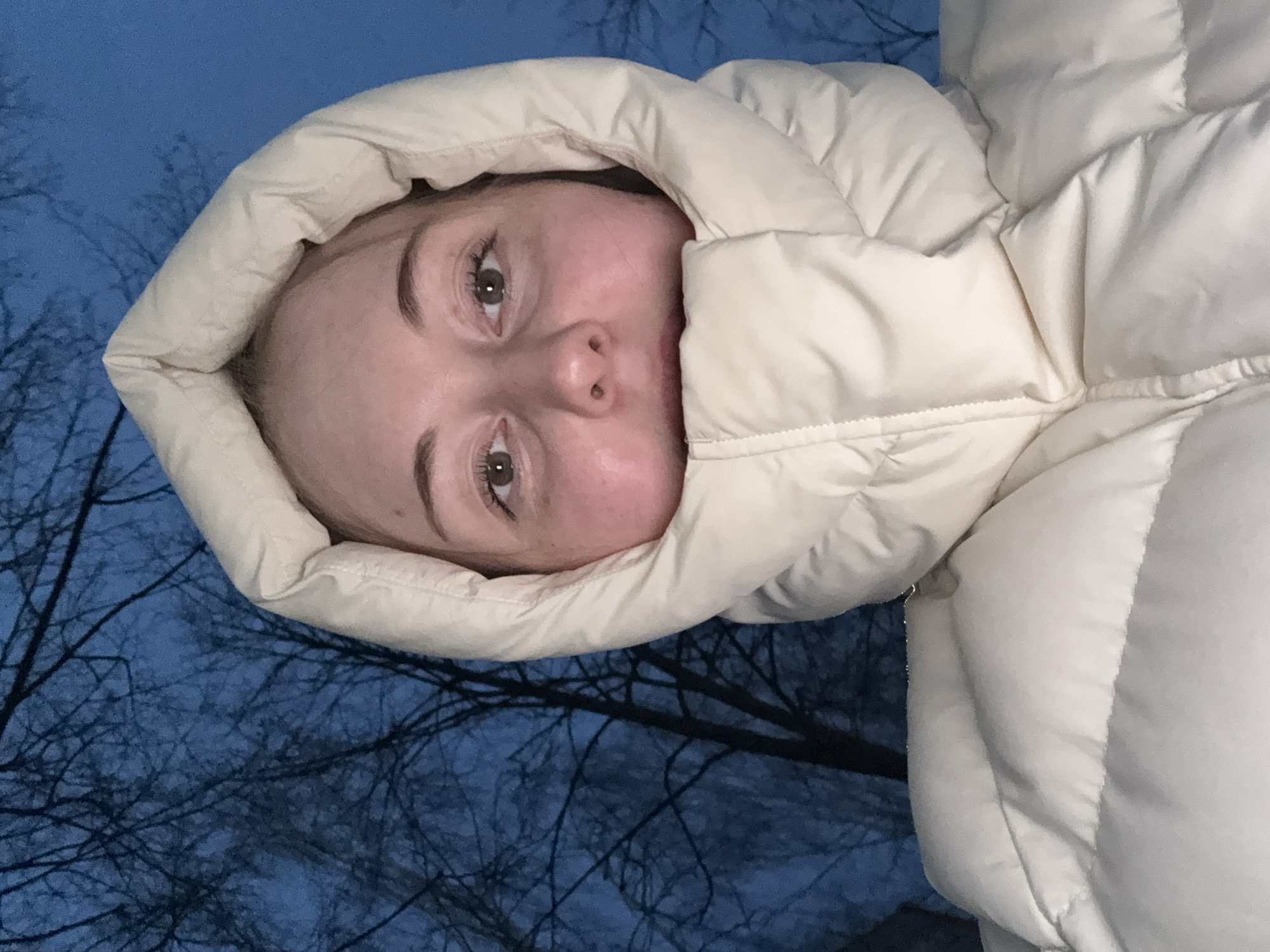 A single individual, appearing female, is depicted in an outdoor setting in the city of Unknown, Unknown. The subject is centered and faces directly into the camera. She is wearing a cream-colored, heavily insulated puffer jacket with the hood pulled up and cinched tightly around her face, covering her ears, neck, chin, and mouth. Her visible facial features include open, dark brown eyes and light skin. The background consists of a dark blue sky, indicative of low light conditions such as dusk, dawn, or night, against which numerous bare tree branches are silhouetted. No specific actions or interactions are evident beyond posing for the image.
