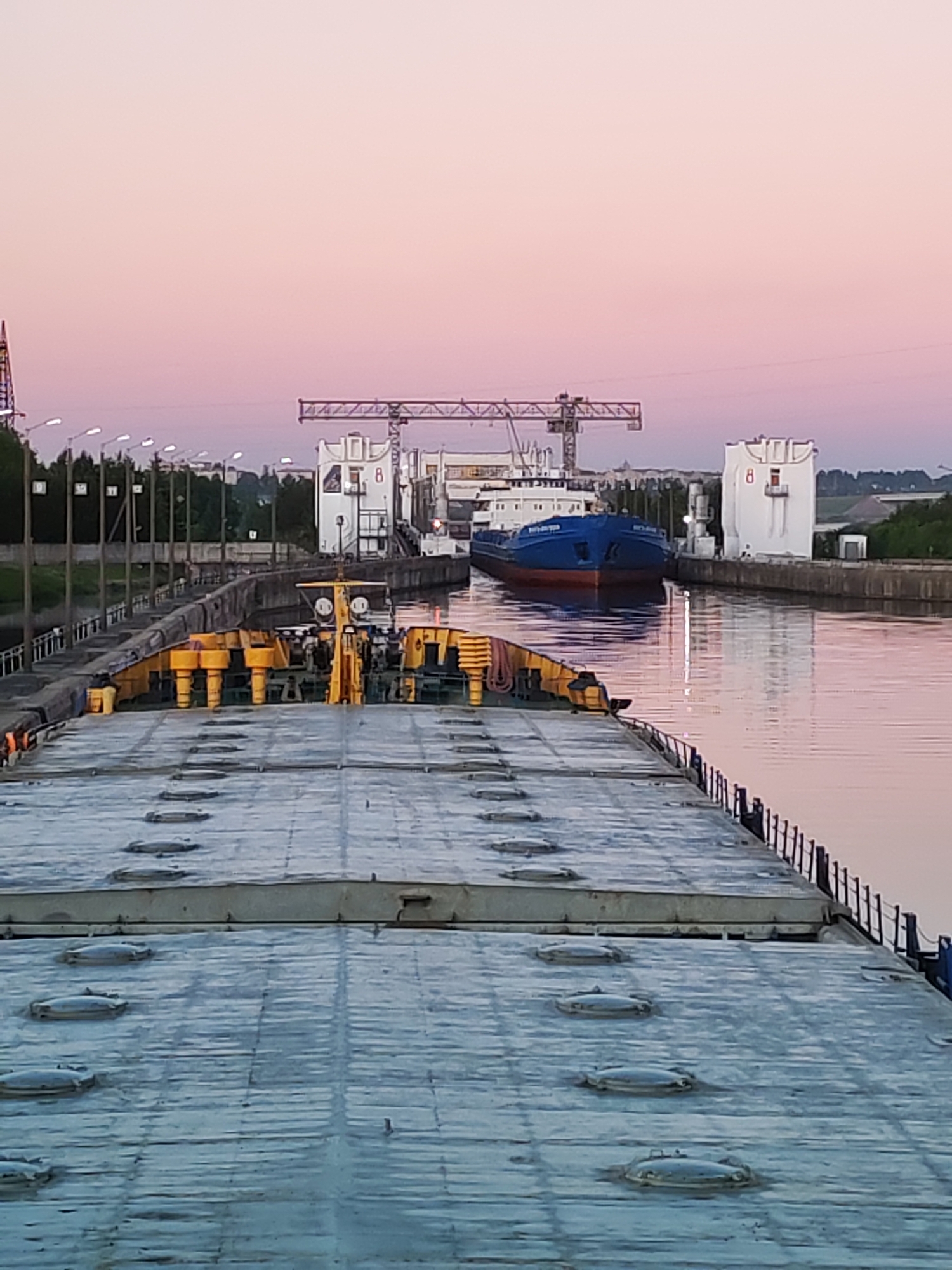 The image captures a detailed view of a canal lock system at either dawn or dusk, characterized by a soft pink and orange gradient in the sky.
In the immediate foreground, the grey, flat deck of a large cargo vessel, possibly a barge, occupies the lower half of the frame. This deck is textured with what appear to be longitudinal seams and features several regularly spaced, oval-shaped metal hatch covers. Near the top edge of this deck, a cluster of yellow-painted mooring bollards and other mechanical components is visible. A dark metal railing runs along the right side of the deck, extending towards the midground.
The vessel in the foreground is positioned within a canal or lock, with its deck appearing slightly wet or reflective. Beyond this vessel, the canal waters are calm, reflecting the muted colors of the sky and the surrounding structures.
In the midground, a blue-hulled cargo ship with a red lower hull and white superstructure is situated within a lock chamber, facing the foreground vessel. Its name, though partially visible, is illegible.
Flanking the lock chamber are two large, white, utilitarian buildings. The building on the right prominently displays a red numeral '8' on its facade, along with a small red door or window and a small balcony. A white vertical cylindrical structure stands next to it. Further back and spanning across the canal, a large industrial gantry crane with a lattice structure is visible, connecting the two sides of the waterway.
The left bank of the canal features a concrete or stone wall, with numerous streetlights on tall poles extending into the distance. Behind these lights, a dense line of green trees is present. In the far background, across the flat horizon, distant fields or low hills can be seen under the pastel sky. No individuals are discernible in the scene. The location is an unidentified canal lock system.