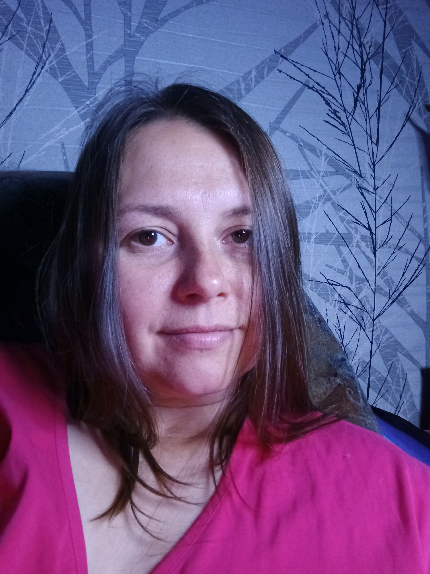 The image is a selfie of a woman with medium-length brown hair, light skin, and brown eyes. She is wearing a bright pink top. She is indoors, and the background appears to be a wall with a patterned wallpaper featuring stylized tree branches. The lighting is moderate, and the image is slightly grainy. No specific location within Novosibirsk, Russia can be determined from the image.
