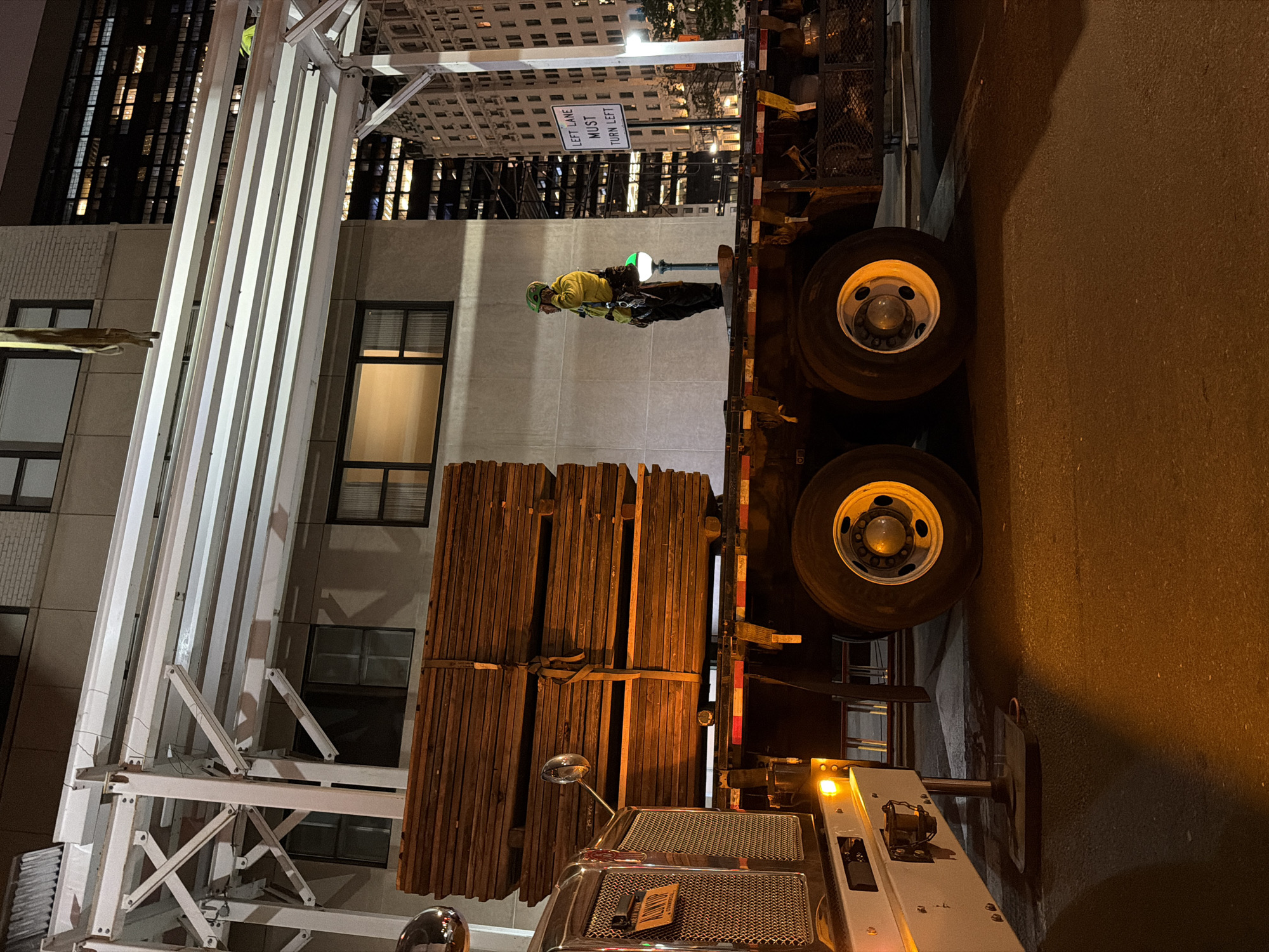 This is a low-angle, medium shot of a flatbed truck loaded with stacked wooden planks. The truck is parked on a city street at night, possibly in New York City, as suggested by a license plate that appears to read "New York". A construction worker wearing a yellow vest and a green hard hat stands on the back of the truck, facing away from the camera and appearing to be talking on a phone. Behind the truck, a building with multiple lit windows is visible, and a street sign reads "LEFT LANE MUST TURN LEFT." The scene is lit by streetlights and the headlights of the truck, casting long shadows on the asphalt.
