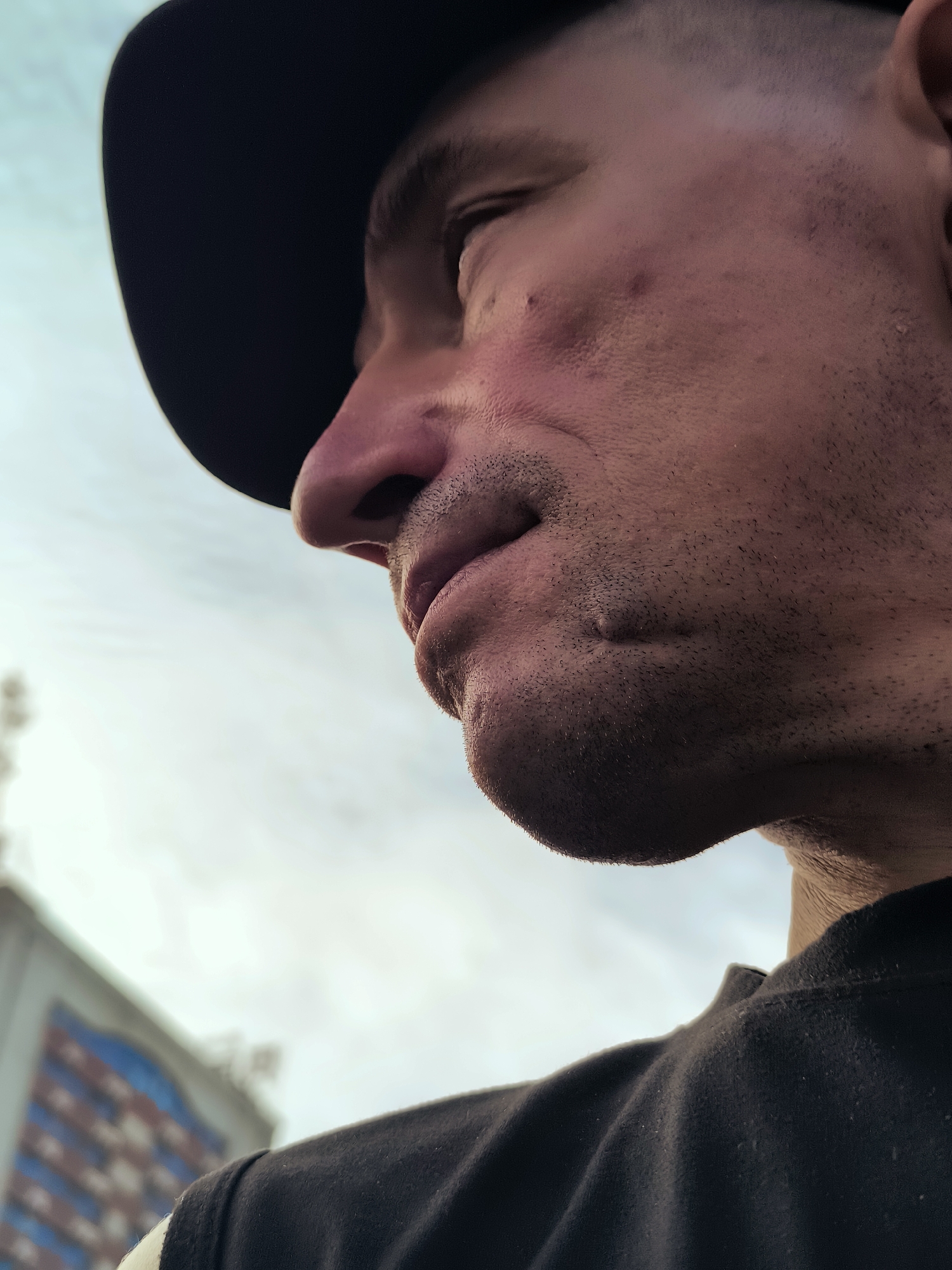 A close-up, low-angle shot shows the profile of a person's face, from the nose down to the chin, wearing a dark baseball cap. The person has stubble on their chin and appears to have some acne on their cheek. The background is a bright, slightly hazy sky with a blurred building visible in the lower left corner. The building has a colorful, patterned facade, suggesting it could be a commercial or residential structure. The scene appears to be outdoors, with the lighting suggesting it might be daytime. The individual's gaze is directed away from the camera, and their expression is unreadable due to the partial view. The image does not provide any specific clues to determine the location within Ürümqi, China.